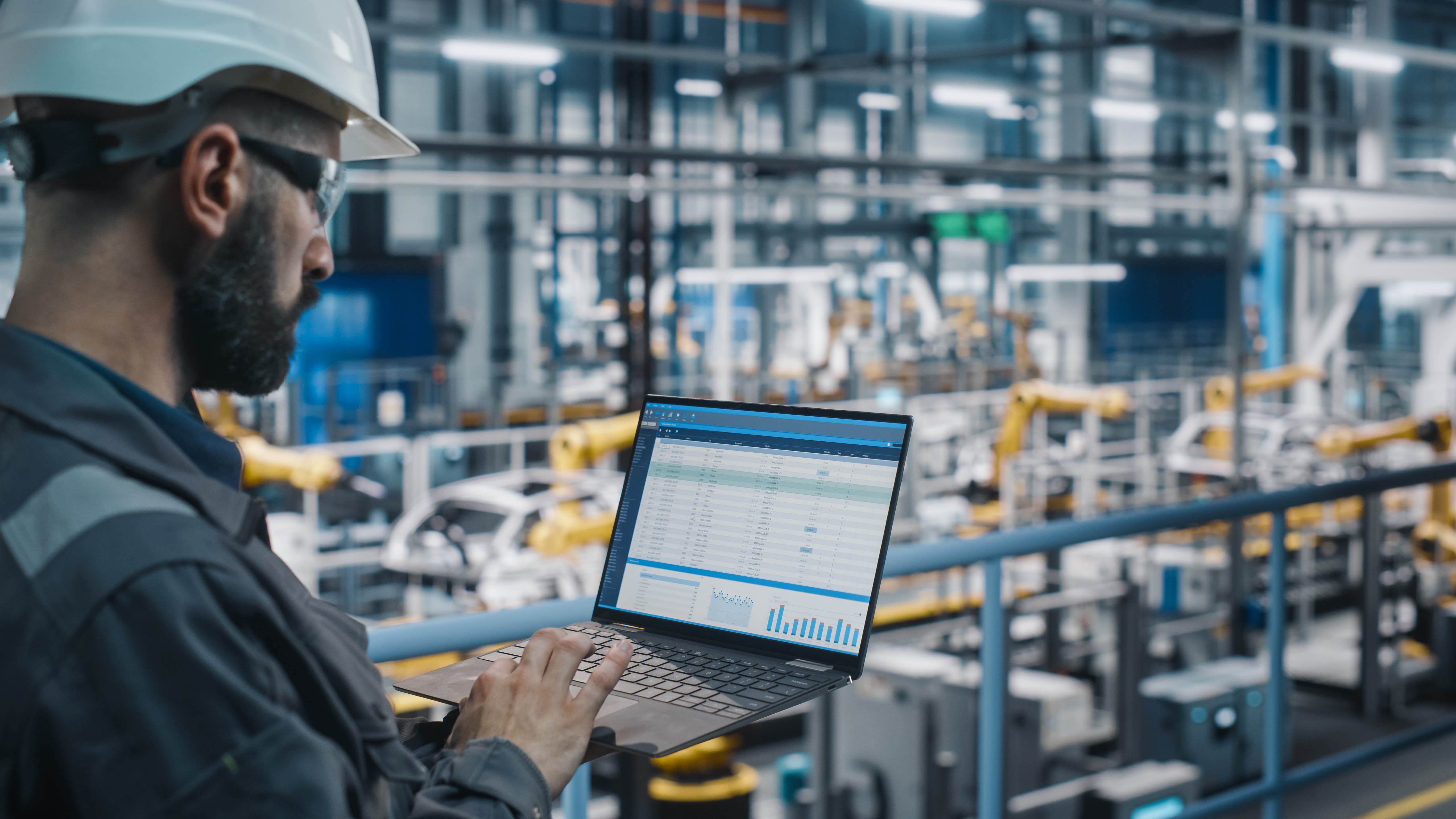 A male worker in PPE at an industrial worksite used his laptop to assess crew management on his worsite.