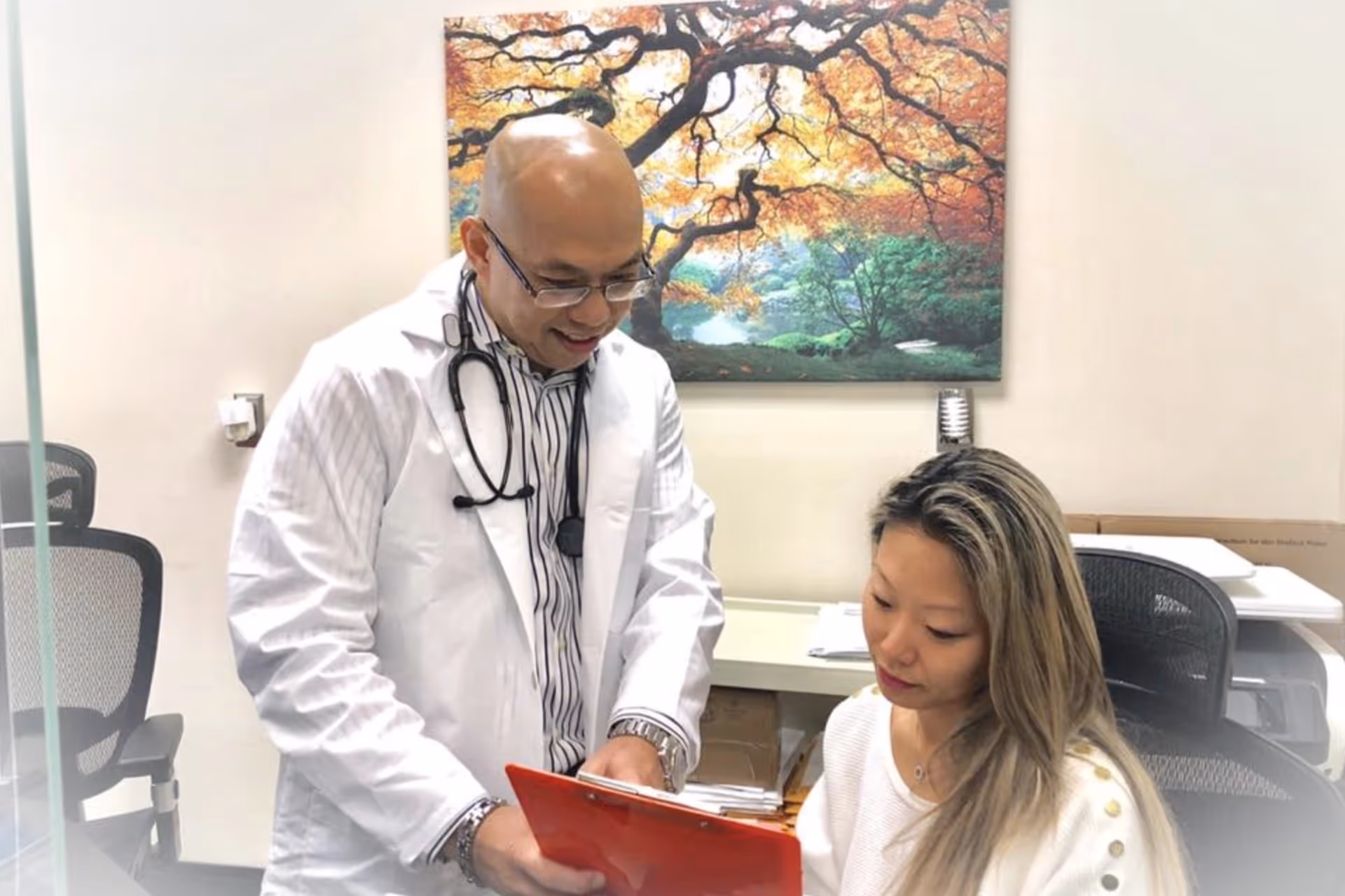 Doctor in white coat showing a red clipboard to a seated woman in a medical office.