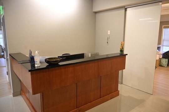 Reception desk with wooden base and black countertop, featuring a computer monitor, hand sanitizer, and a decorative bowl.