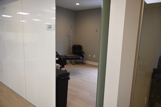 Open hospital exam room with black chairs, gray walls, light wooden floor, and medical equipment stand.
