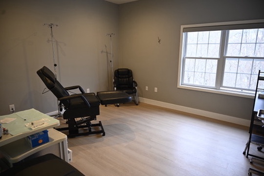 Minimalist medical examination room with two black chairs, an examination table, IV stand, and a large window on a beige wall.
