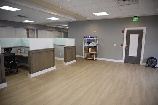 Modern office space with light wood flooring, two desks with frosted glass partitions, a door with a glass panel, and shelving unit with office supplies.