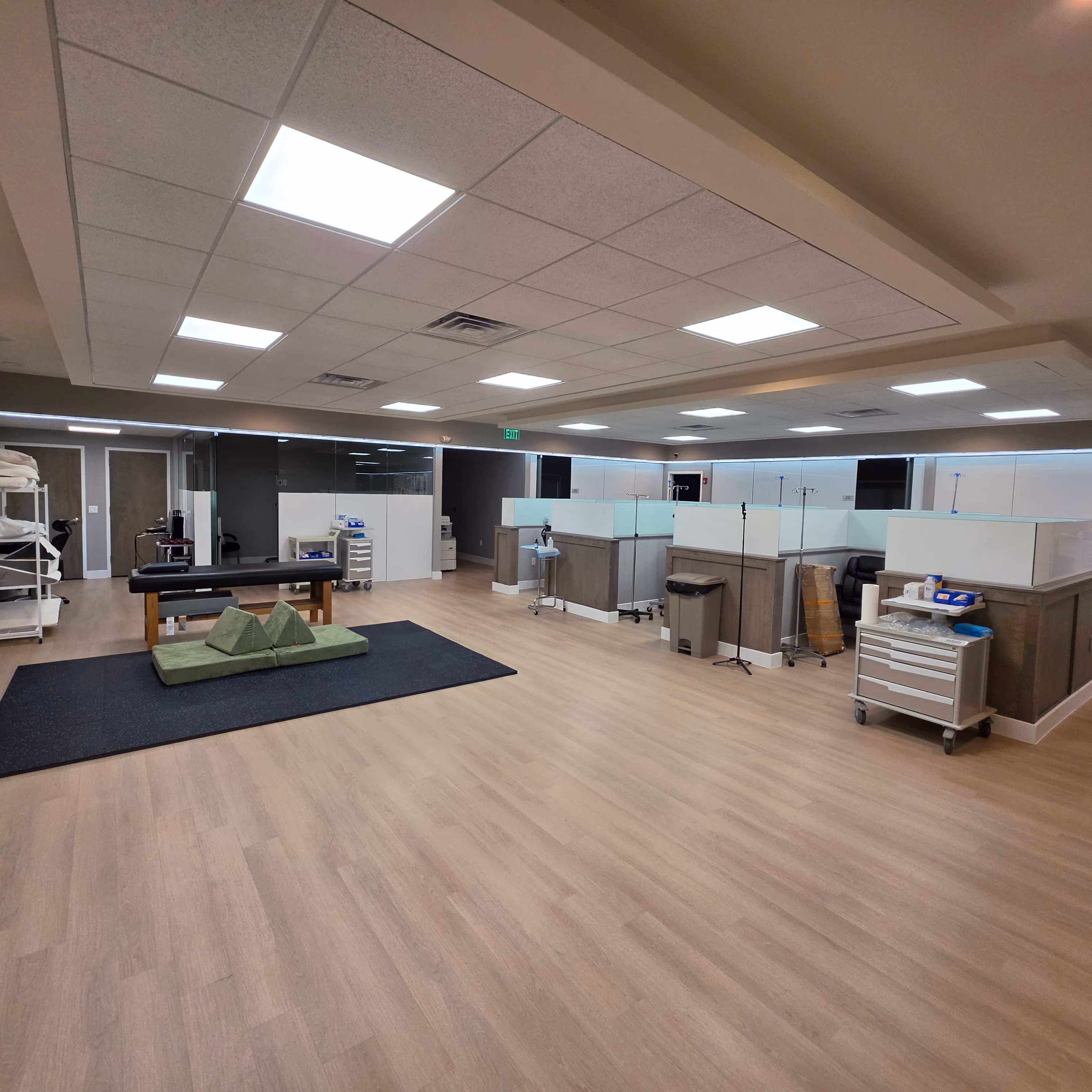 Modern office space with light wood flooring, two desks with frosted glass partitions, a door with a glass panel, and shelving unit with office supplies.