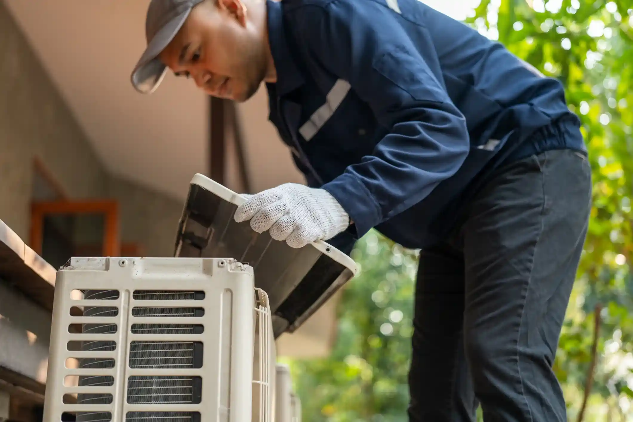 A technician in a blue uniform and cap works on an outdoor air conditioning unit, inspecting it closely.
