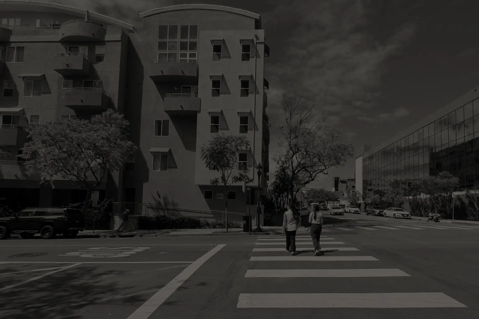 Two people walking on a crosswalk in downtown San Diego with buildings and parked cars around.