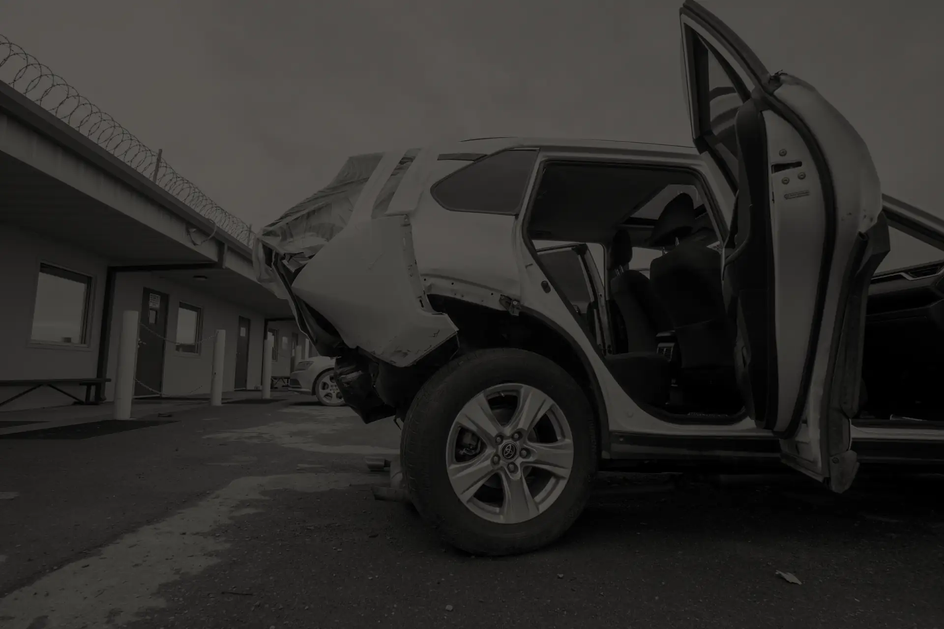 Wrecked SUV with rear damage and open doors parked in a salvage yard near buildings with barbed wire.