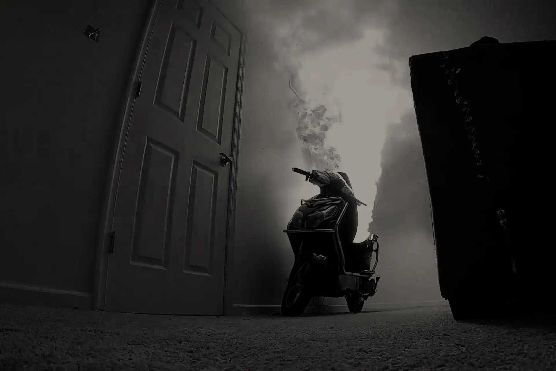 Electric scooter on fire indoors near a door and a large black object in a dimly lit room.