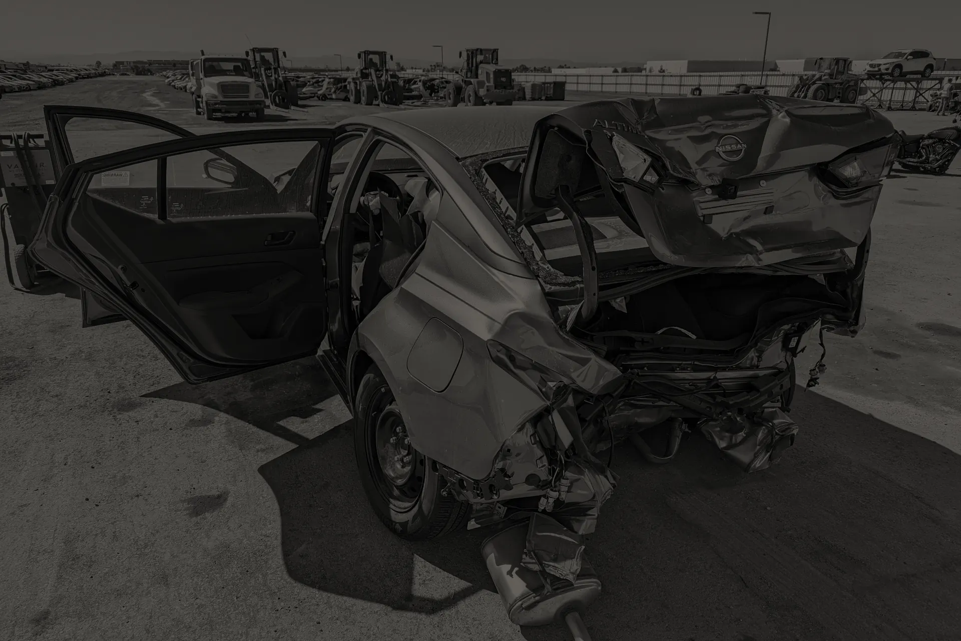 Rear of a severely damaged Nissan Altima with open doors in a lot with vehicles and equipment.