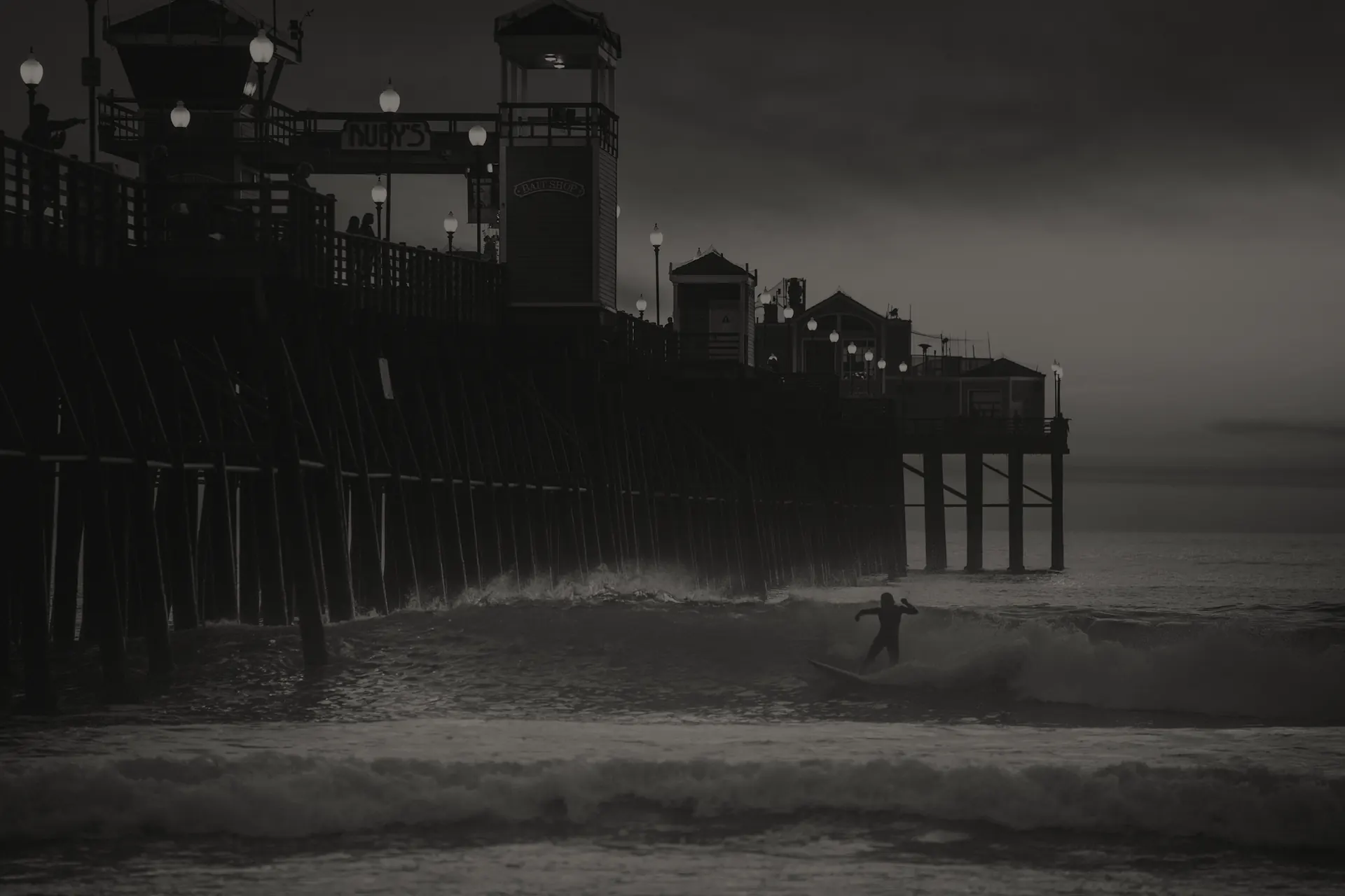Silhouetted surfer riding wave near Oceanside pier at dusk under cloudy skies.
