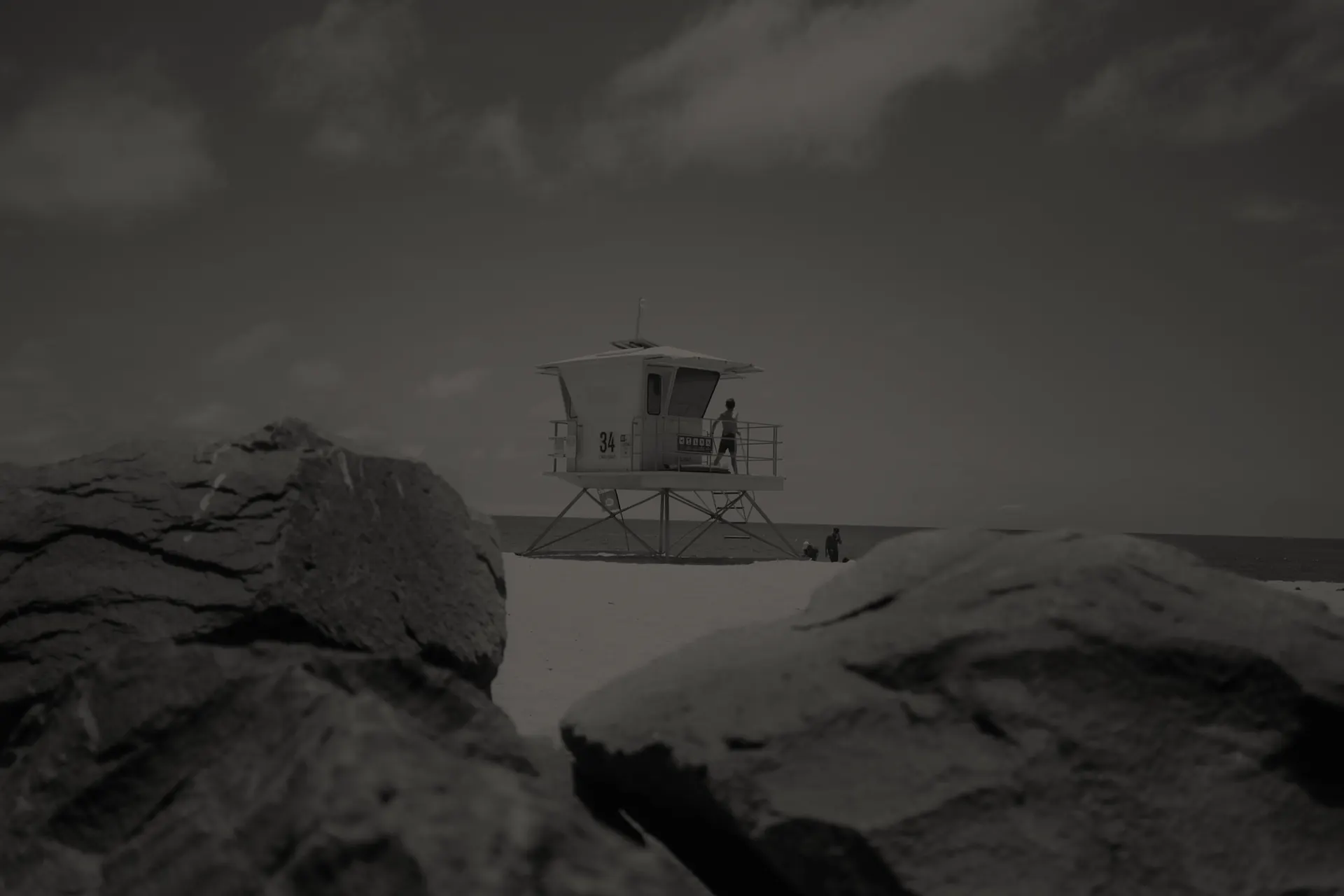 Lifeguard tower 34 on a sandy Carlsbad beach with rocks in the foreground and a person standing on the tower.