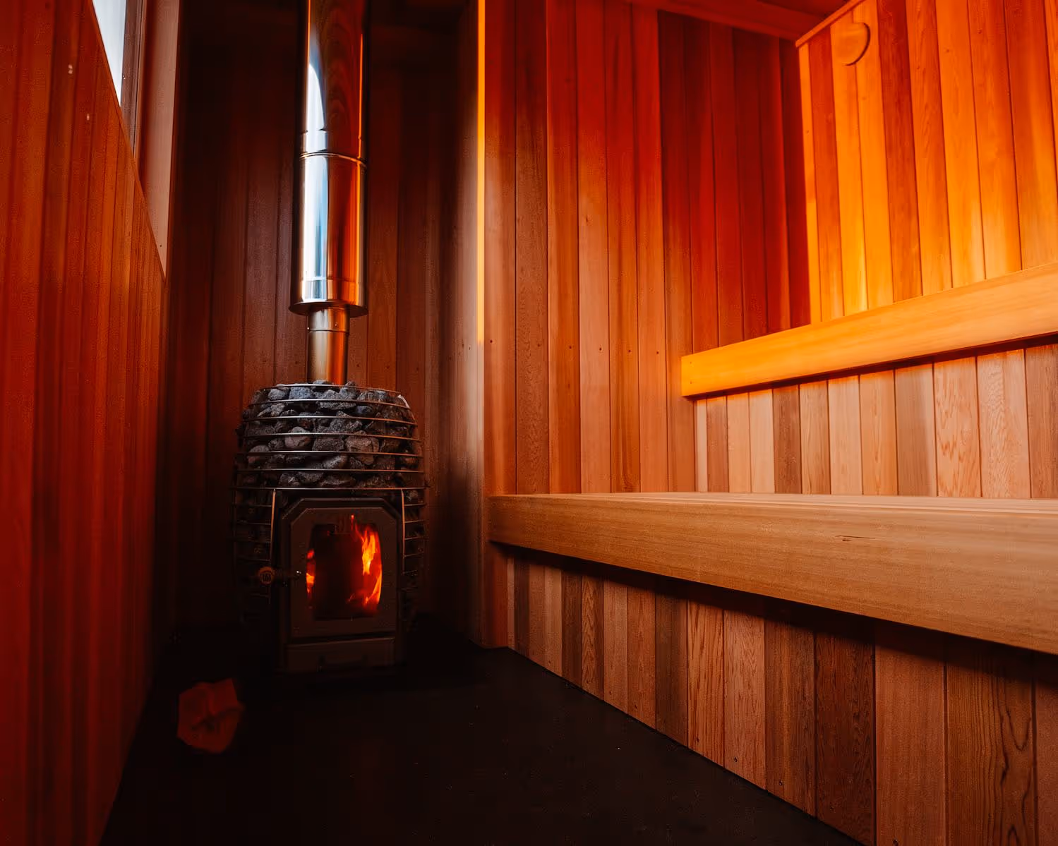 Interior view of wooden sauna showing black sauna stove with glowing flames and metal chimney pipe, surrounded by warm wood paneling on walls and ceiling with tiered wooden benches.