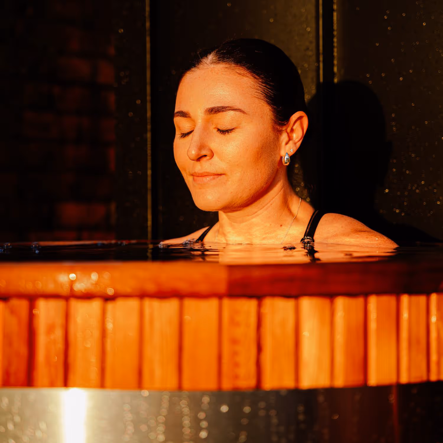 Woman with eyes closed in a wooden ice plunge tub with warm golden lighting, water surface reflecting ambient light in evening setting.