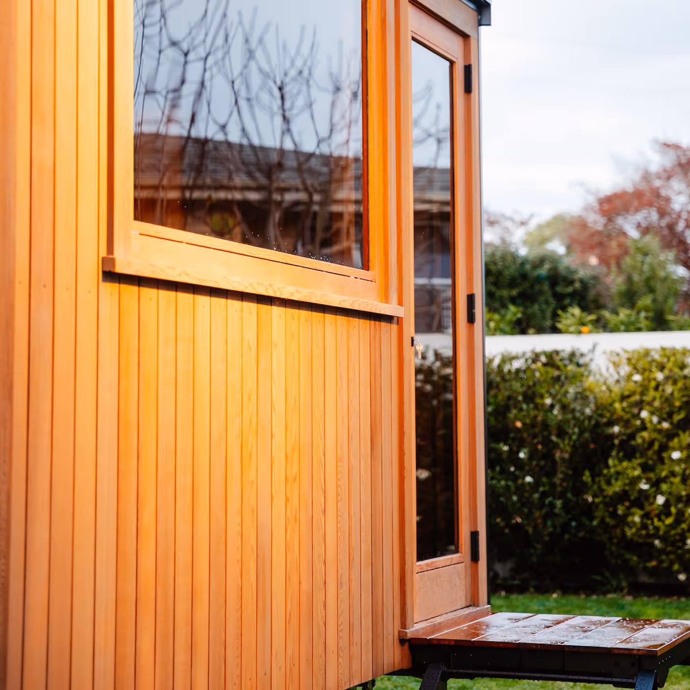 Exterior view of mobile sauna unit with natural wood cladding, large window, and open door revealing wooden interior, set against green lawn and garden backdrop.