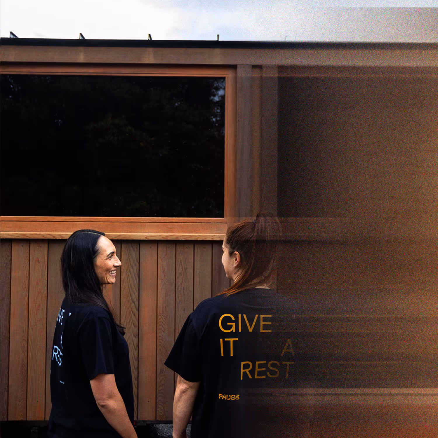 Lauren and Maysie in black shirts standing outside the Pause mobile sauna, viewed from behind. Maysies shirt reads "GIVE IT A REST" with "Pause" branding.