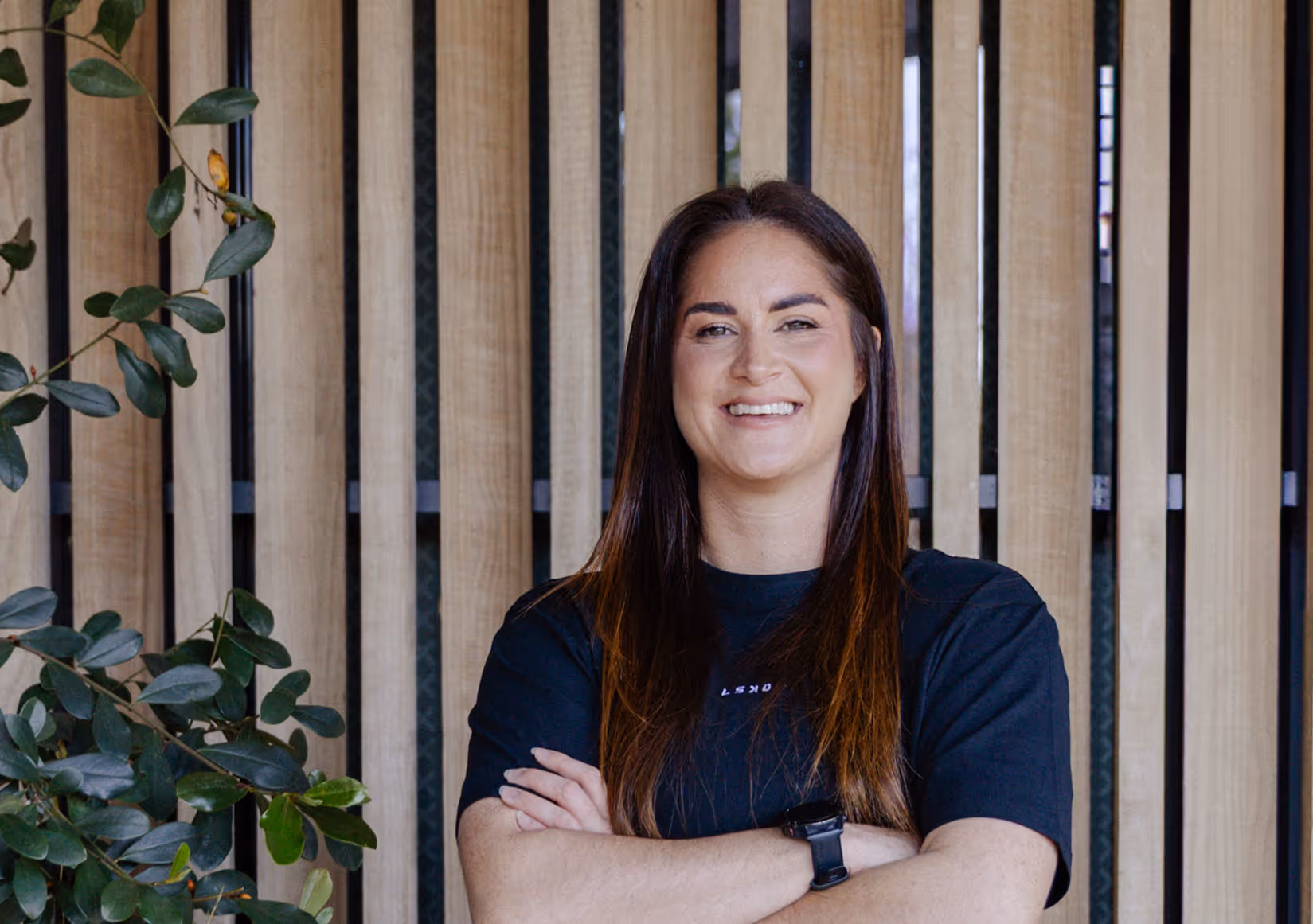 Professional headshot of Maysie smiling, wearing dark top, arms crossed, standing against natural wood slat background with green plant visible.