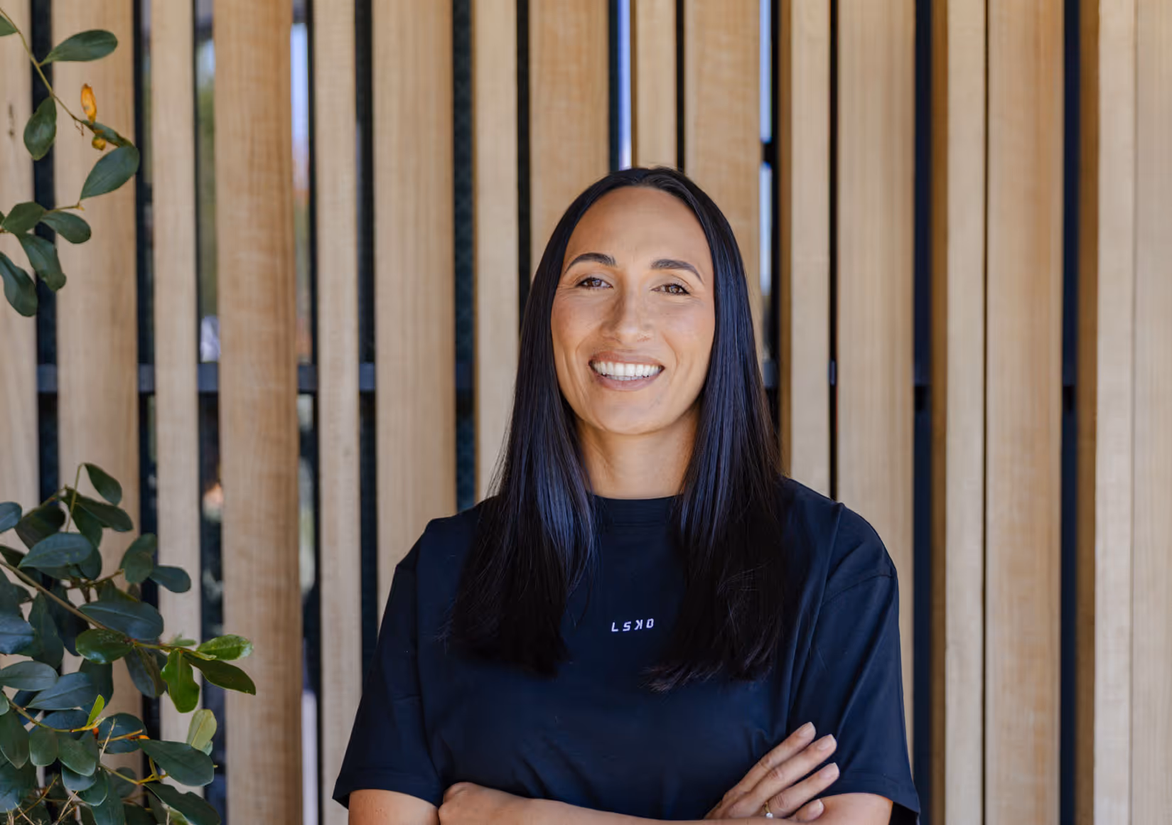 Professional headshot of Lauren smiling, wearing dark top, arms crossed, standing against natural wood slat background with green plant visible.