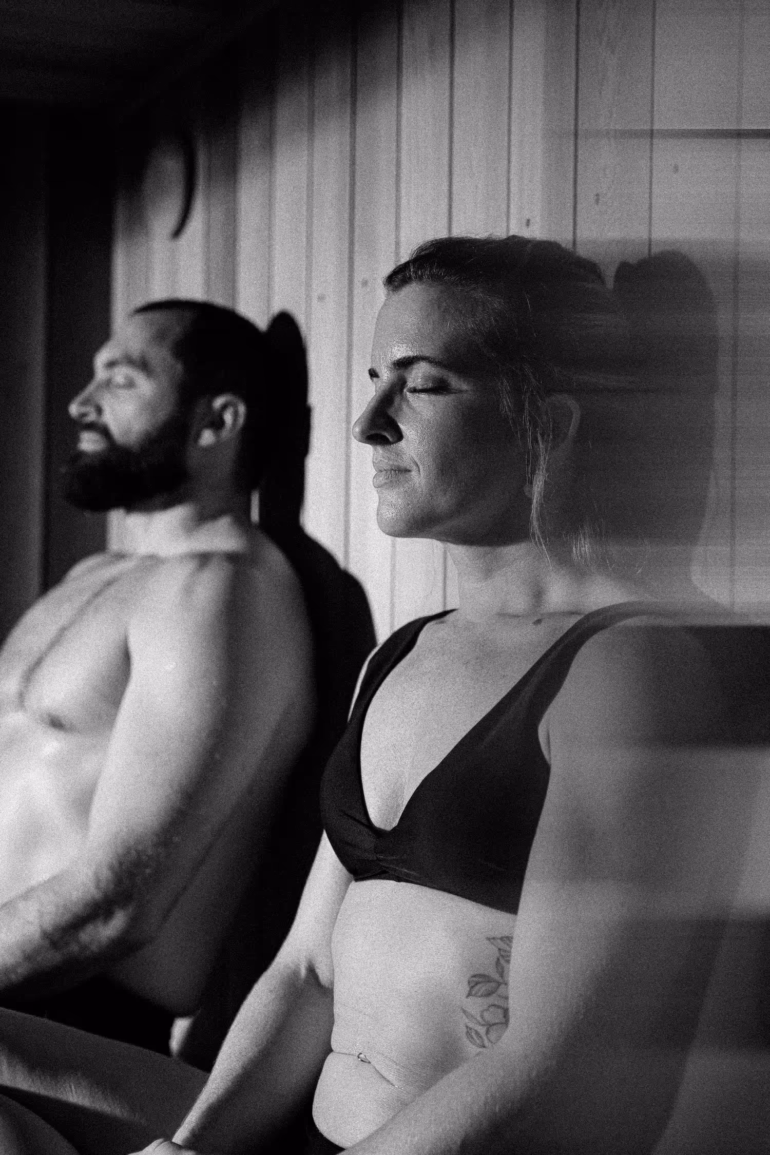 Black and white photograph of man and woman sitting together on wooden sauna bench, both wearing minimal athletic clothing with eyes closed in relaxed poses against vertical wood paneling.