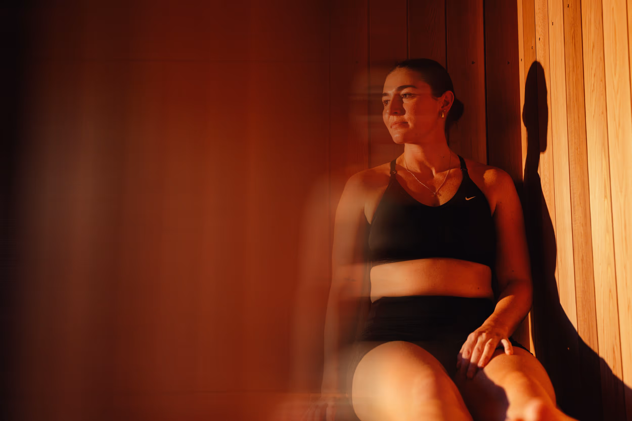 Woman in black sports bra sitting in wooden sauna with warm orange lighting, eyes closed in peaceful meditation leaning against vertical wood paneling.