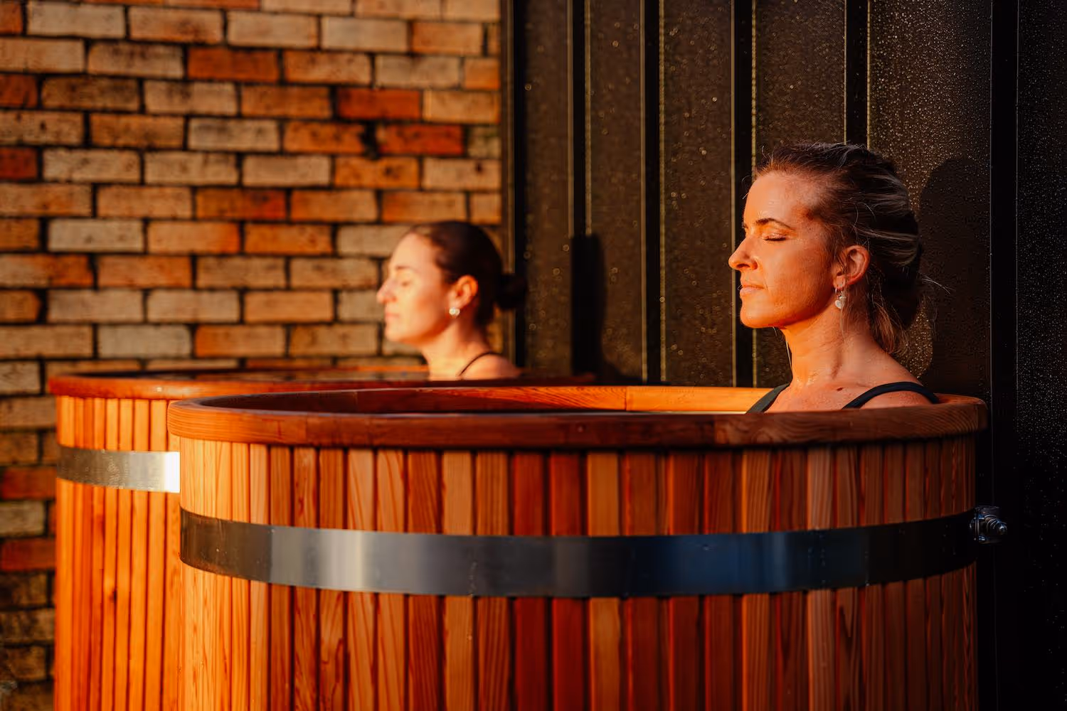 Two women relaxing in circular wooden ice plunge tub with warm lighting, brick wall visible in background, both appearing serene with eyes closed.