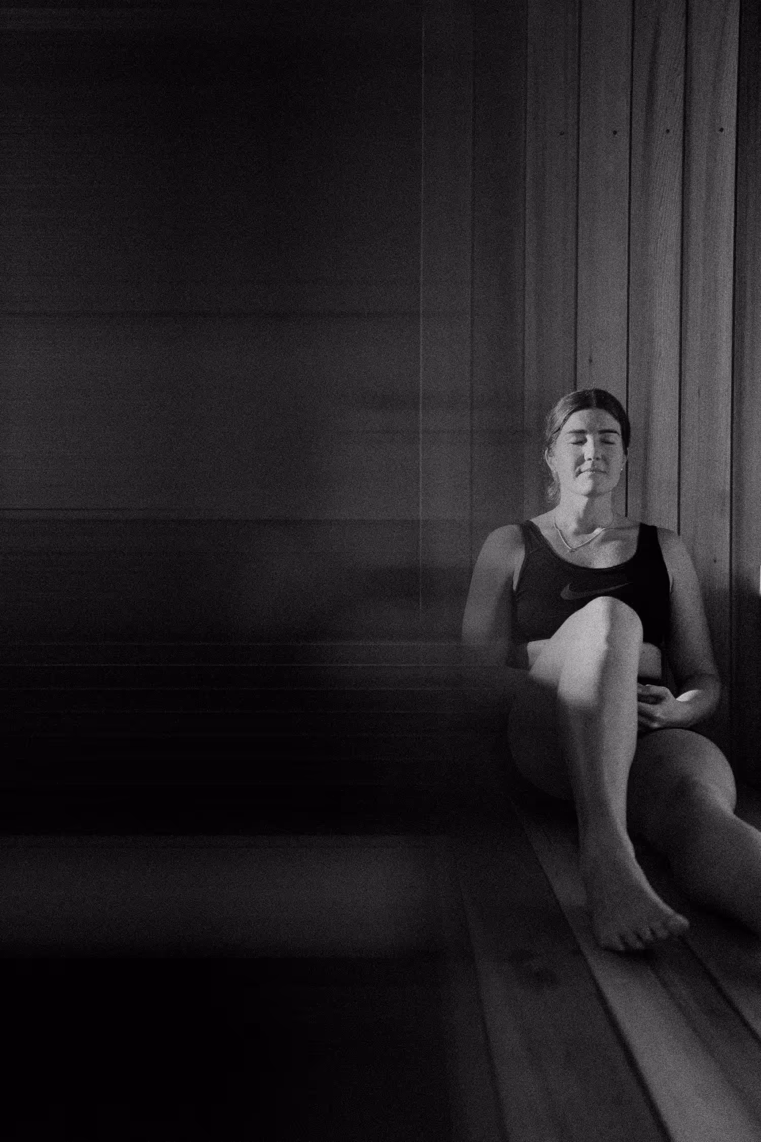 Black and white photograph of woman in dark athletic wear sitting peacefully on wooden sauna bench with eyes closed and hands resting on her knees.