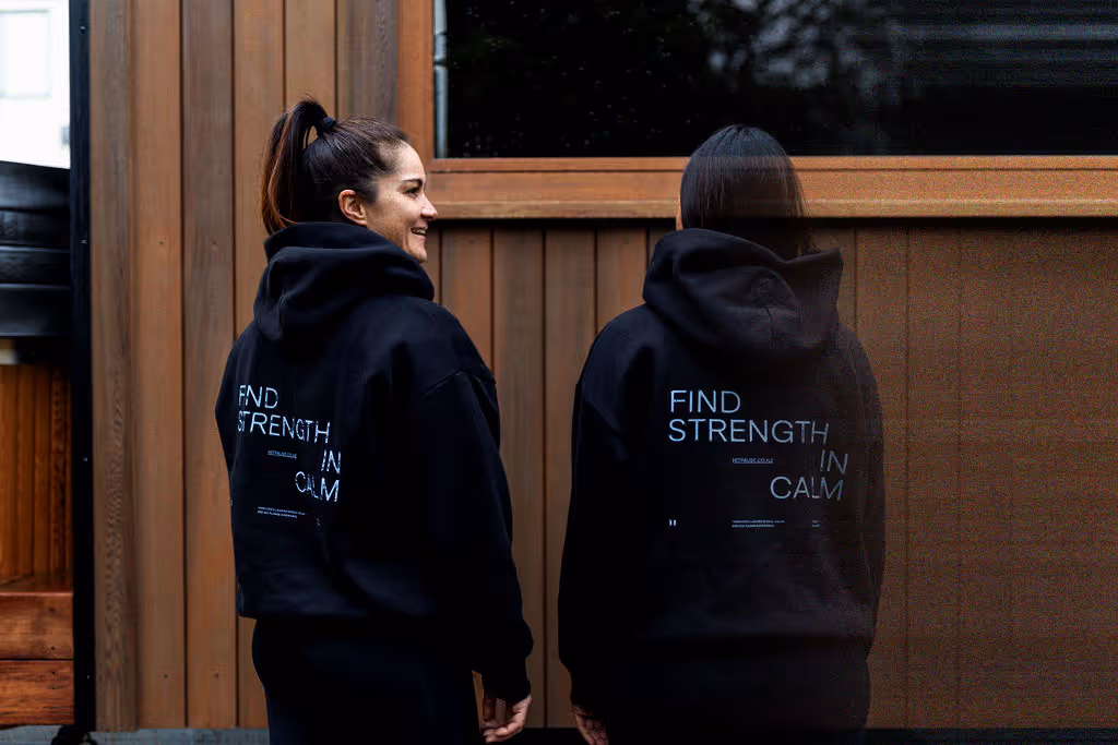 Lauren and Maysie in black hoodies standing outside the Pause mobile sauna, viewed from behind. Hoodies display "FIND STRENGTH IN CALM" text on the back.