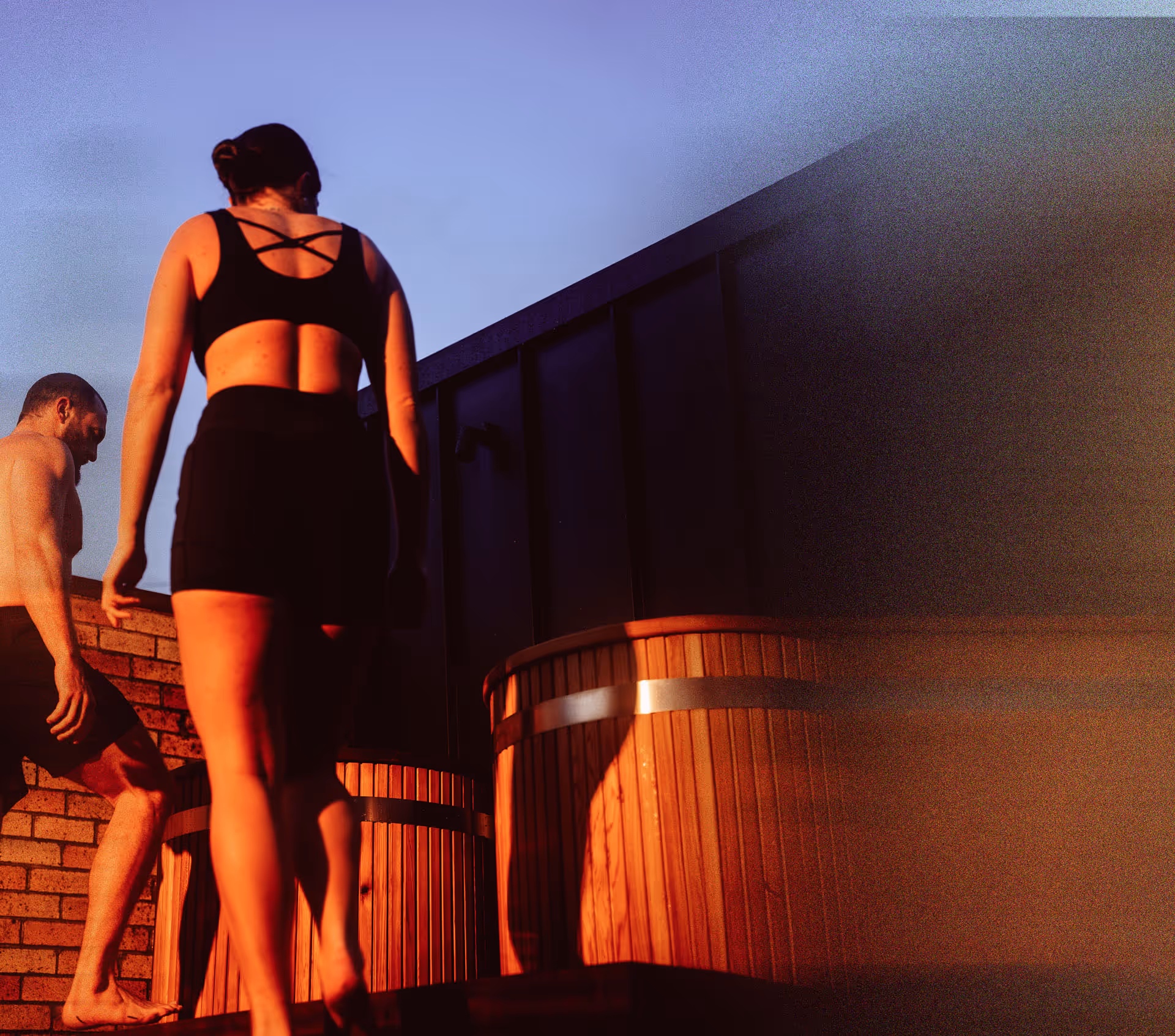 A man and woman on wooden deck beside circular wooden ice plunge tub. The woman is wearing black athletic wear with crossed back straps.