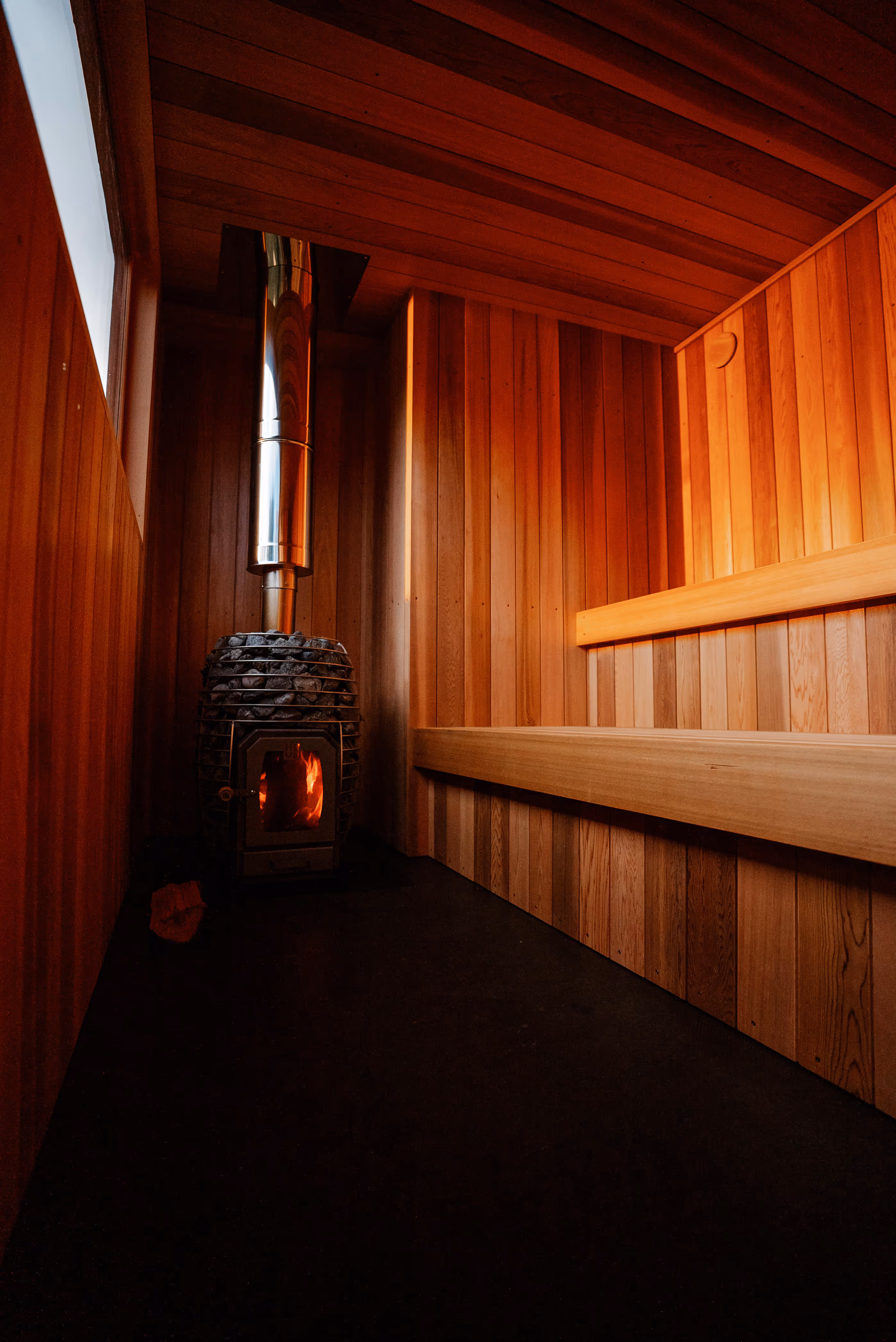Interior view of wooden sauna showing black sauna stove with glowing flames and metal chimney pipe, surrounded by warm wood paneling on walls and ceiling with tiered wooden benches.