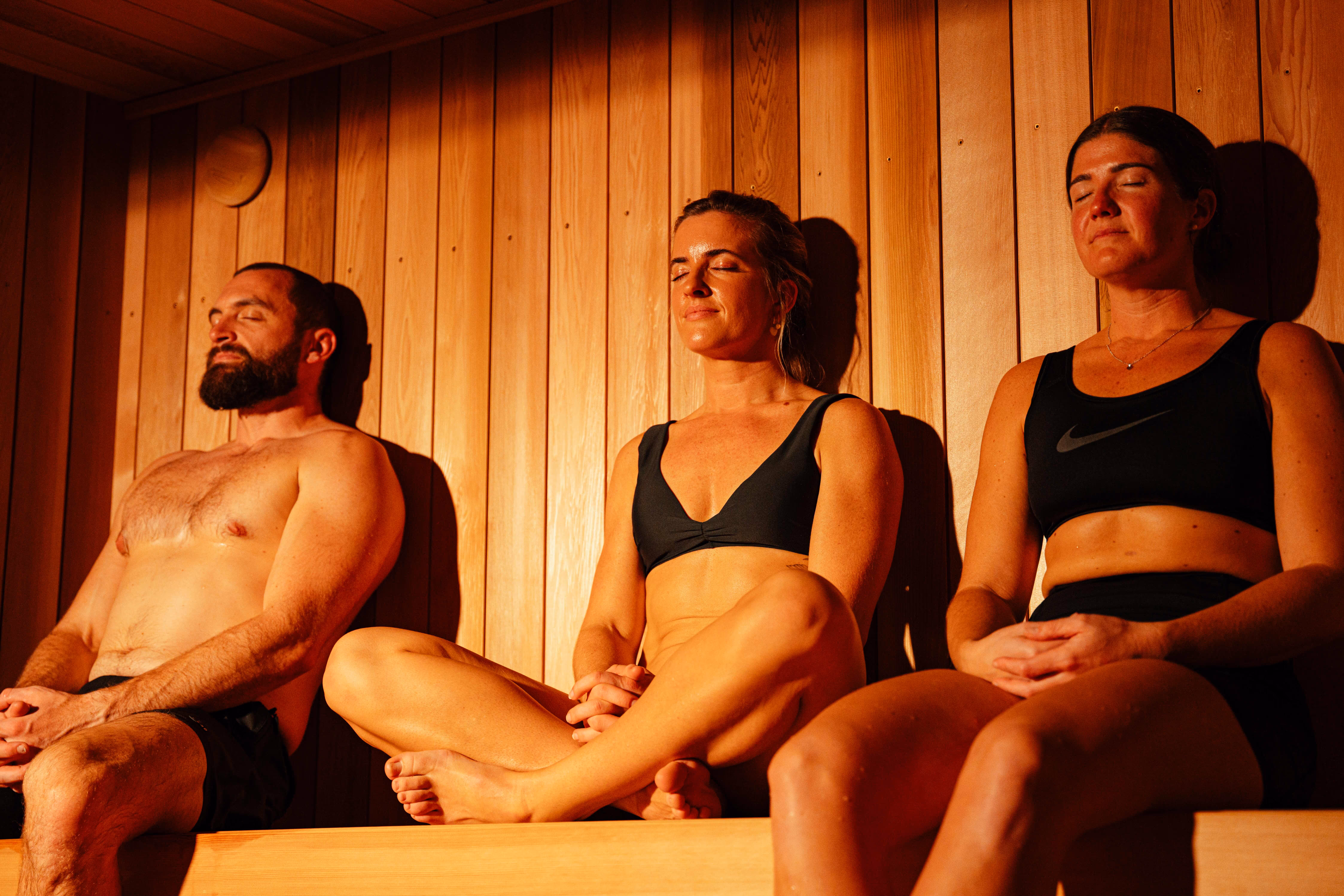 Three people sitting peacefully in wooden sauna with eyes closed in meditation poses - one bearded man and two women in dark athletic wear, all bathed in warm golden lighting against vertical wood paneling.