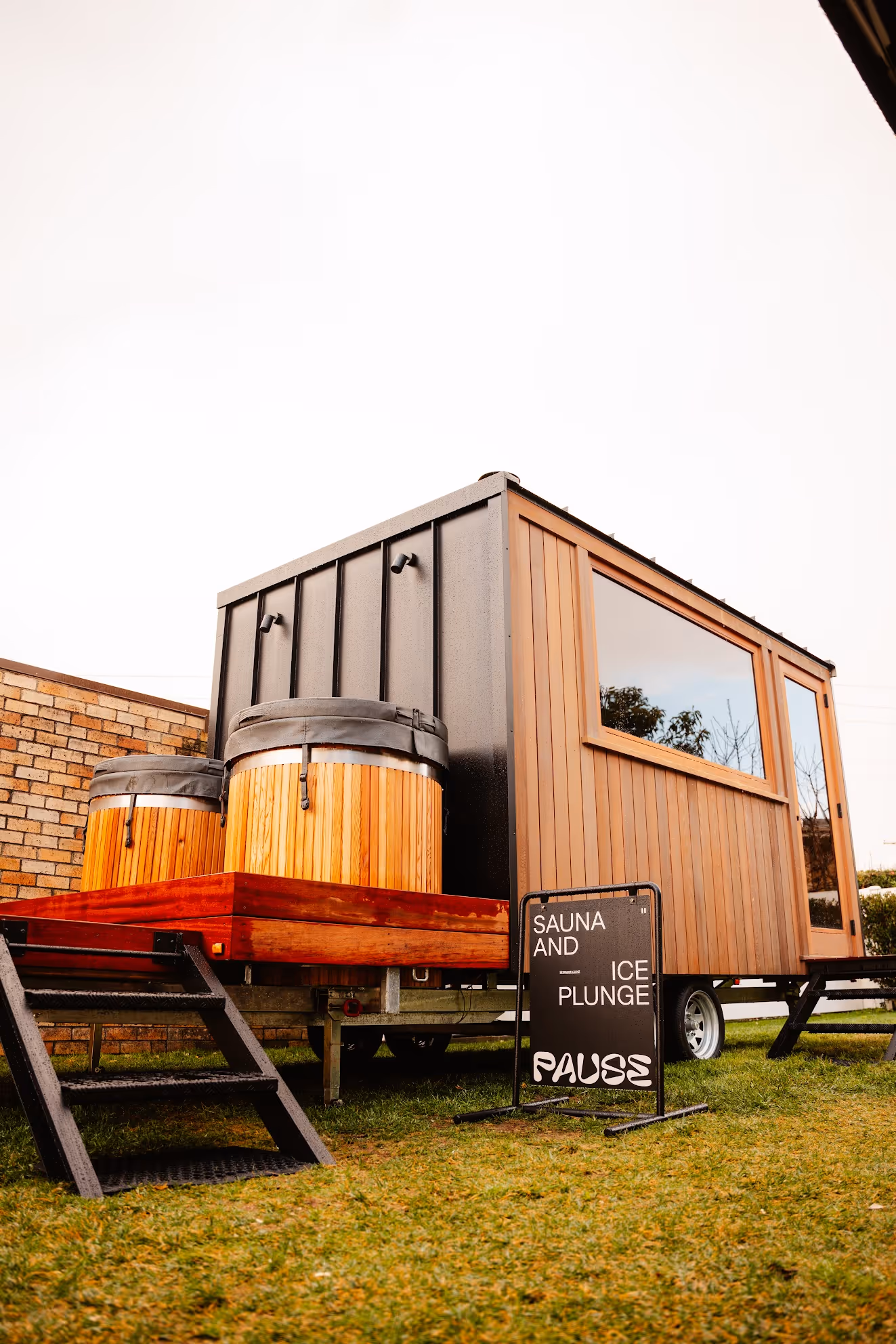 Mobile sauna and ice plunge trailer with wood-clad exterior and large window, featuring two wooden ice plunge barrels on red wooden platform.