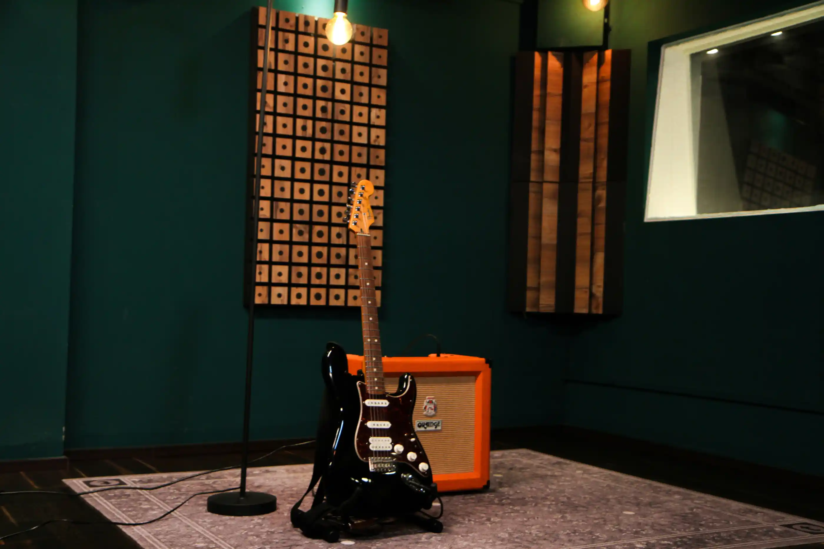 Black electric guitar on a stand in a music studio next to an orange guitar amplifier with acoustic panels on the walls.