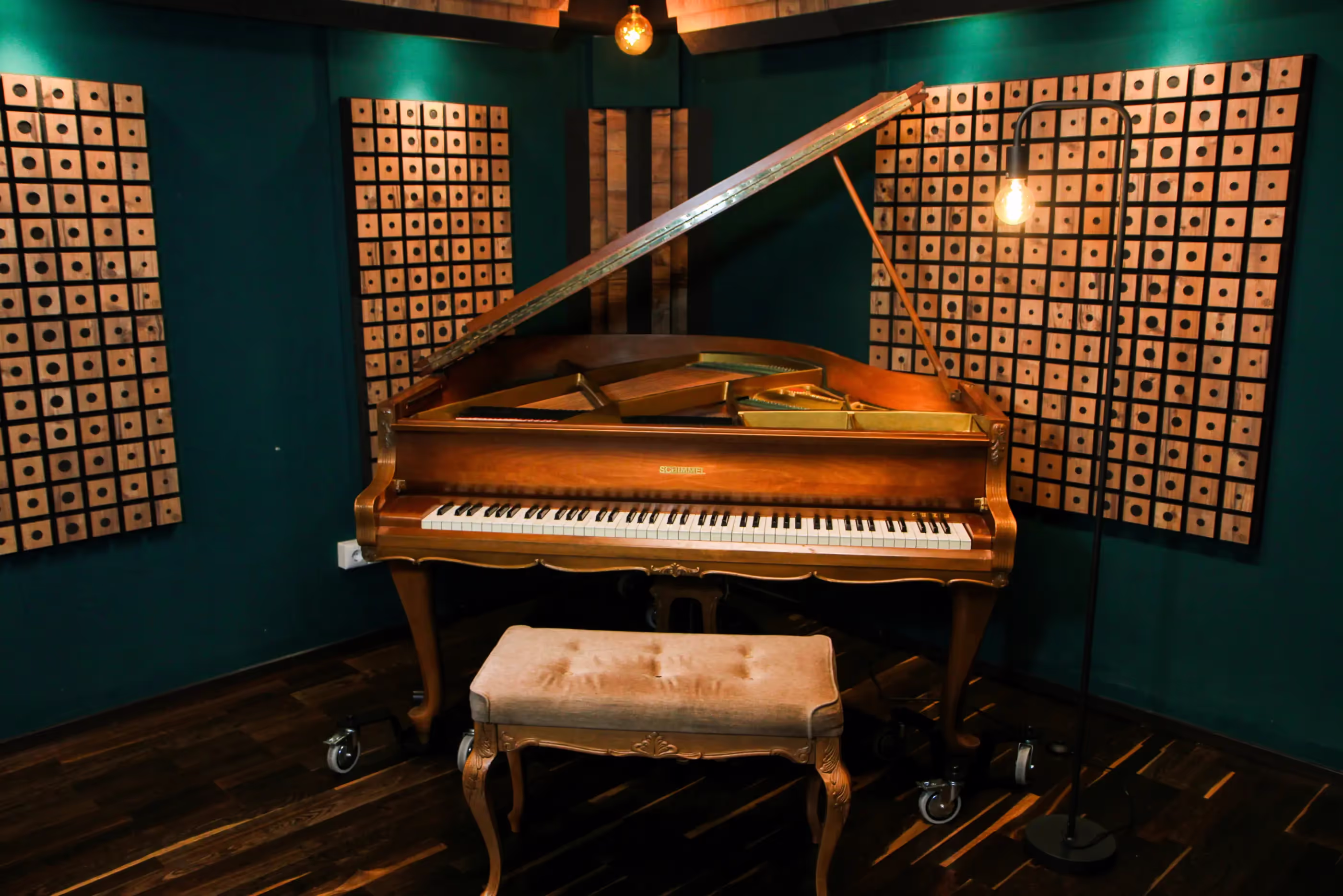 Brown grand piano with an open lid in a recording studio, accompanied by a cushioned bench and a standing lamp with an exposed bulb.