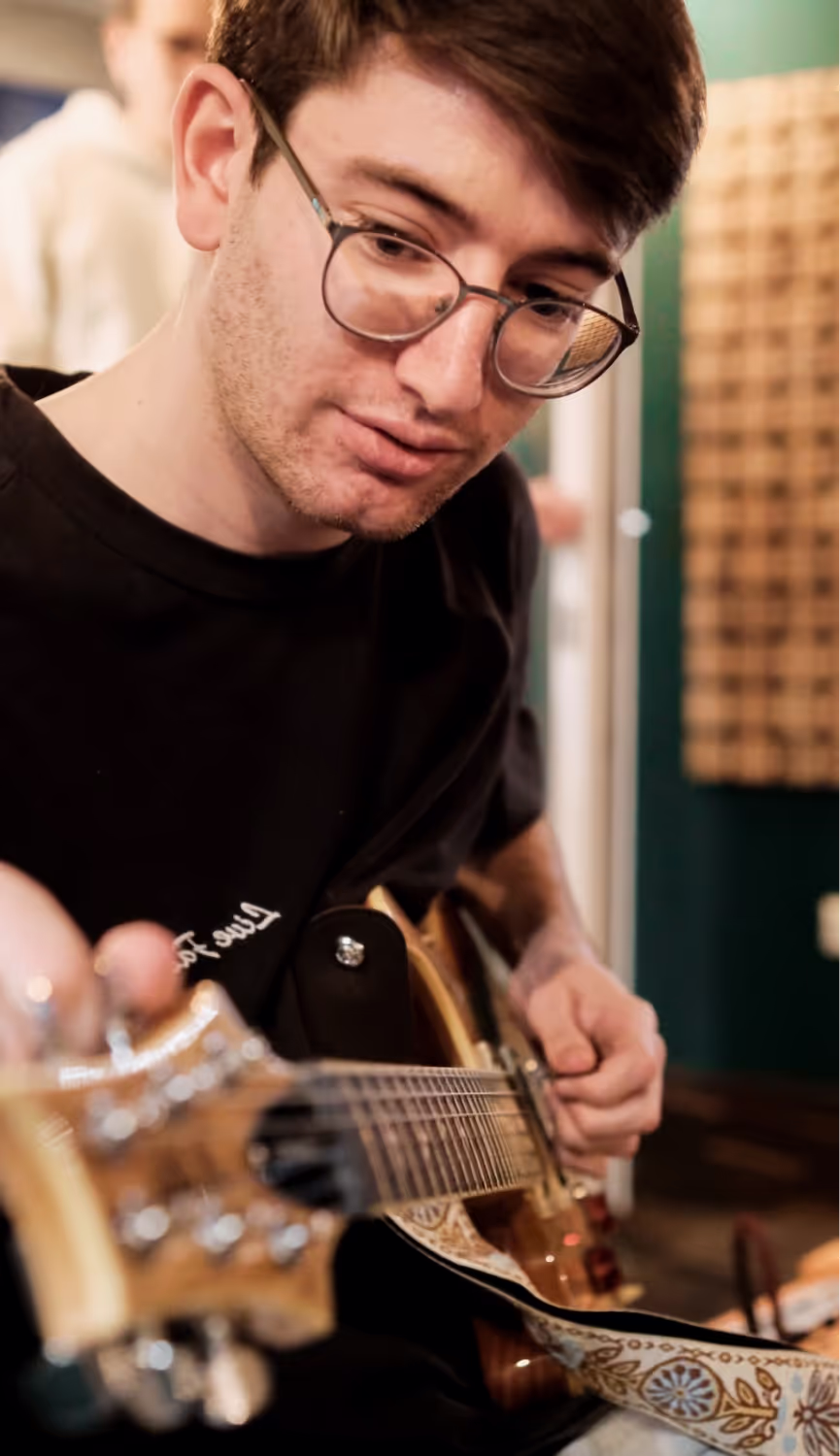 Young man wearing glasses playing an electric guitar with a patterned strap indoors.