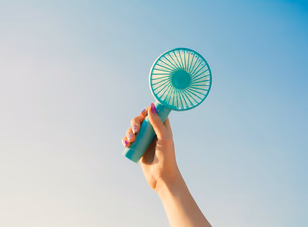 Hand hält türkisen Mini-Ventilator vor blauem Himmel.