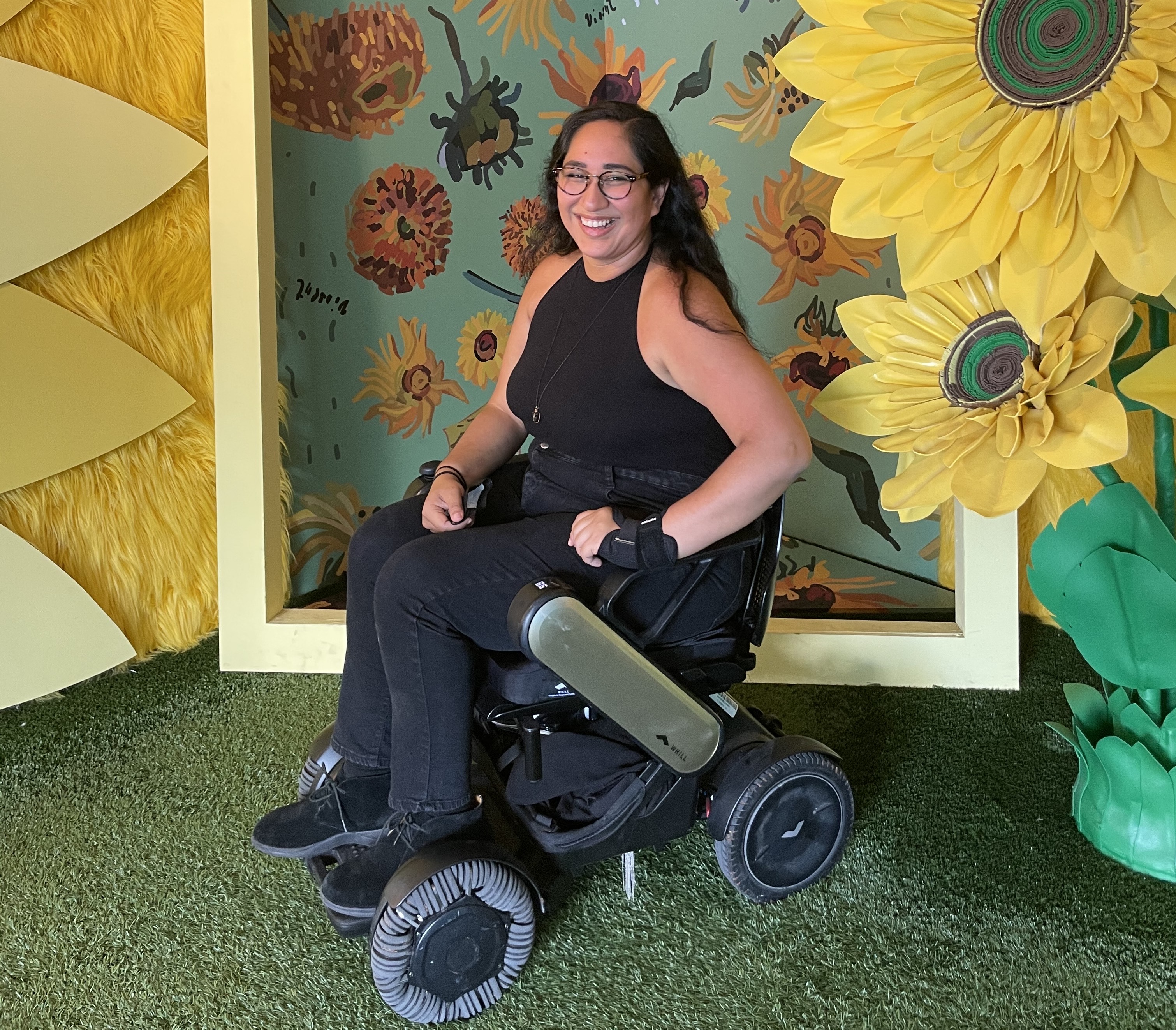 Annie Segarra smiles in a power wheelchair, surrounded by large sunflowers and floral decor.