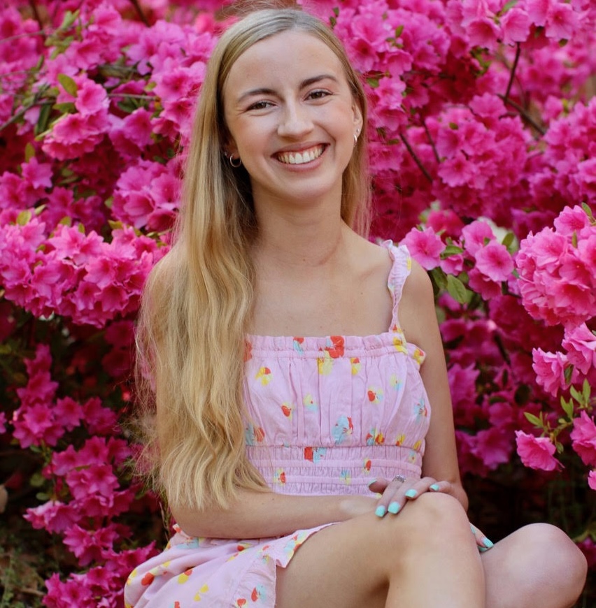 Woman sitting in front of bright pink flowers, smiling and wearing a light pink dress.