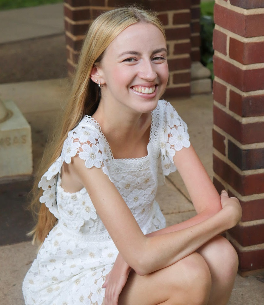 Smiling woman in a white lace dress sitting near a brick wall.