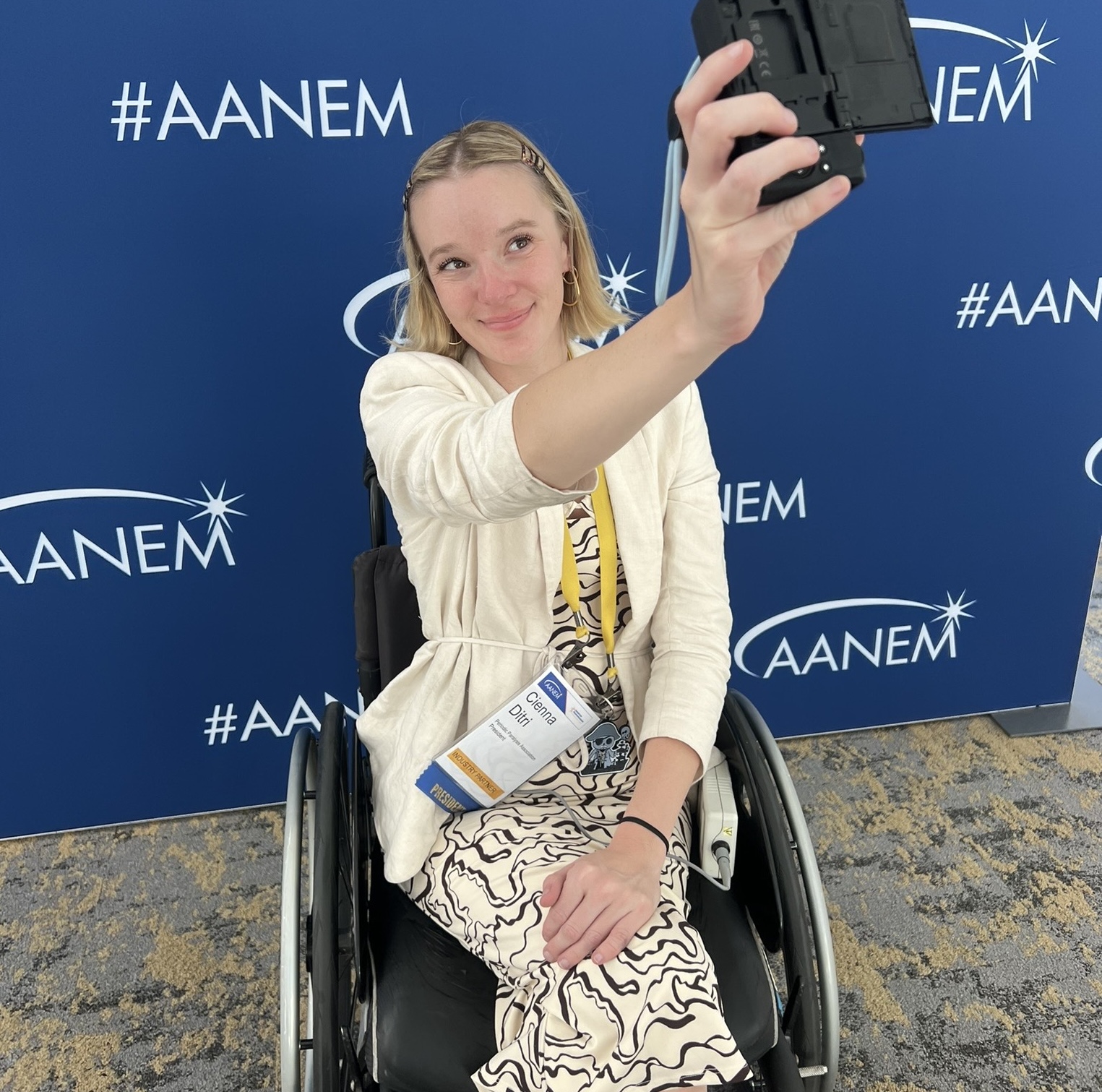 Woman in a wheelchair taking a selfie at a conference backdrop.