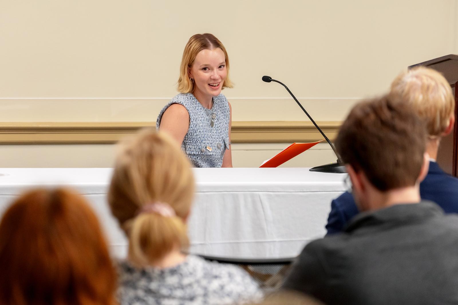 Cienna speaking at a table with a microphone during an event.