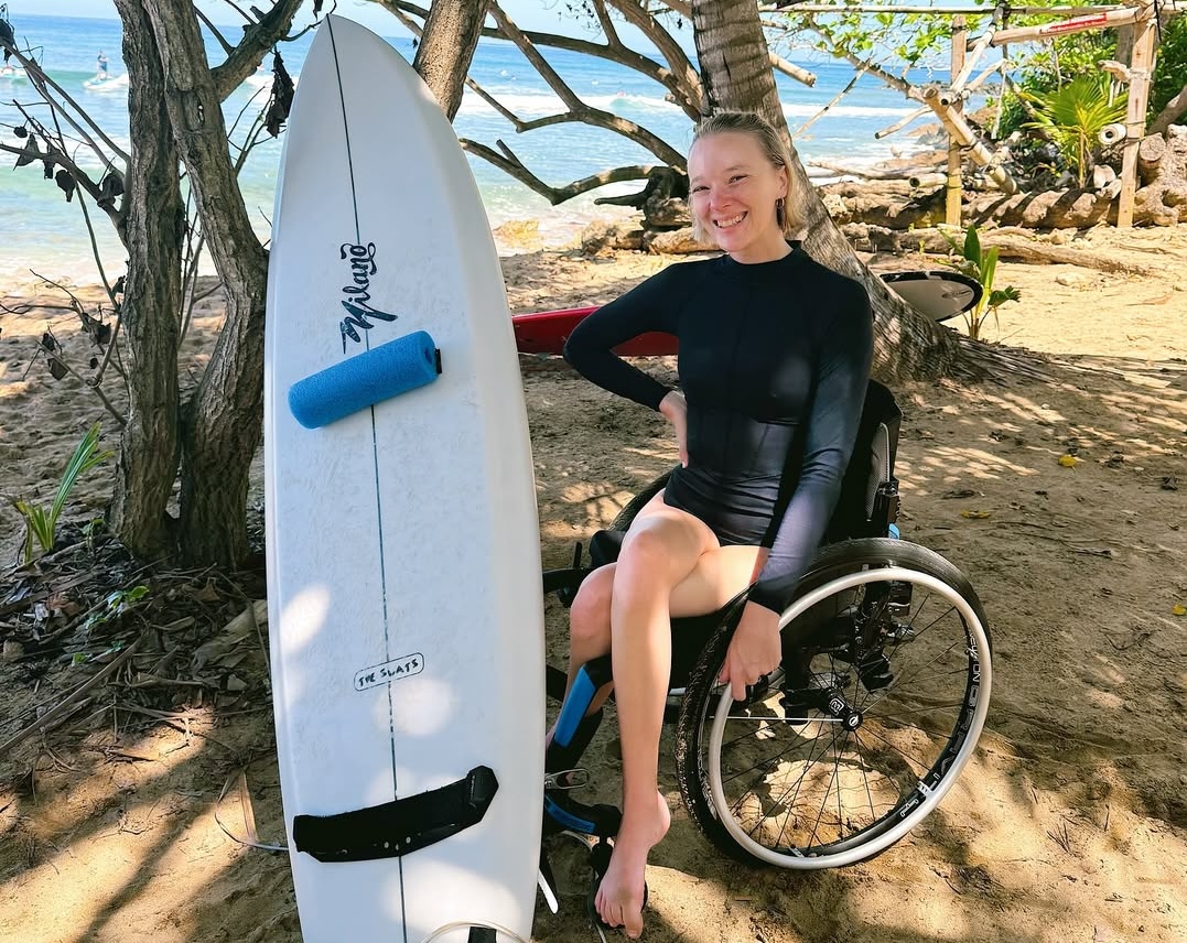 Woman in a wheelchair posing on the beach next to a surfboard.