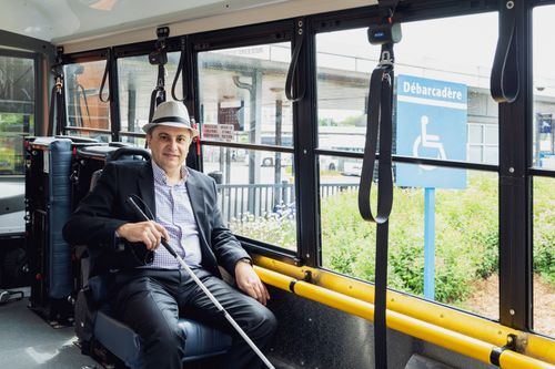 Photo of Dory Jade sitting on an accessible bus, holding a white cane near a disability access sign.