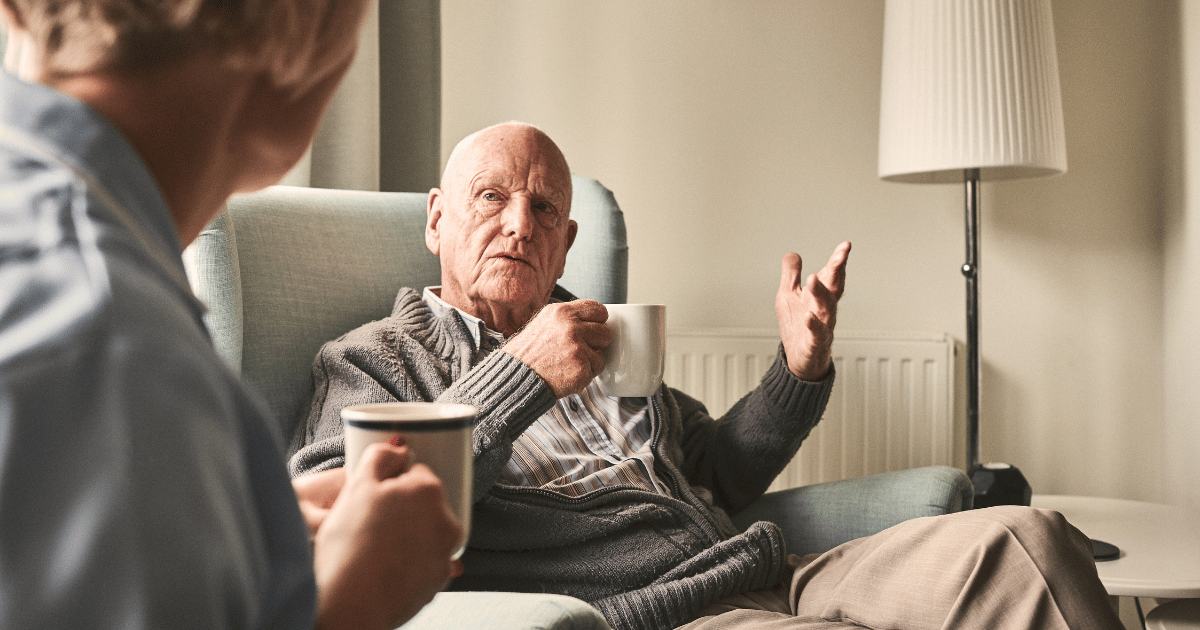 A older man holds a cup of tea whilst talking