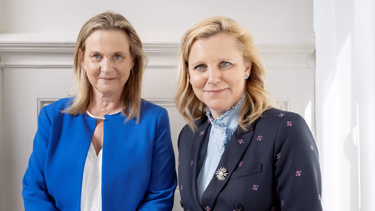 Two professional women wearing formal attire, facing the camera in a well-lit office setting.