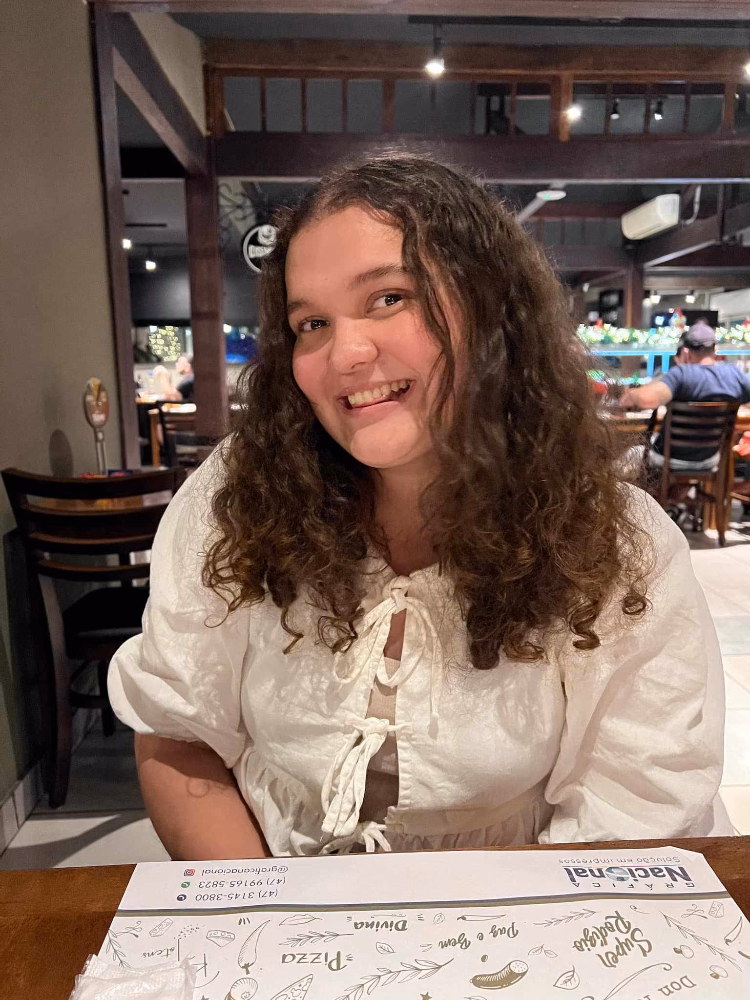 Smilende kvinne med krøllete hår i hvit bluse sitter ved et bord på restaurant.