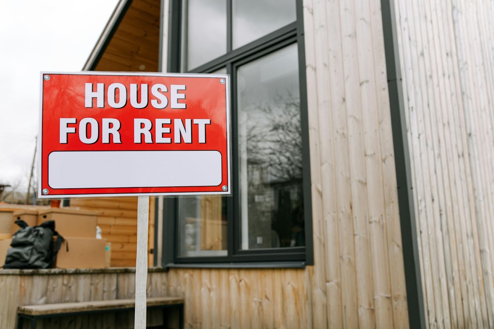A red and white 'House for Rent' sign standing in front of a modern wooden residential building with moving boxes on the porch.