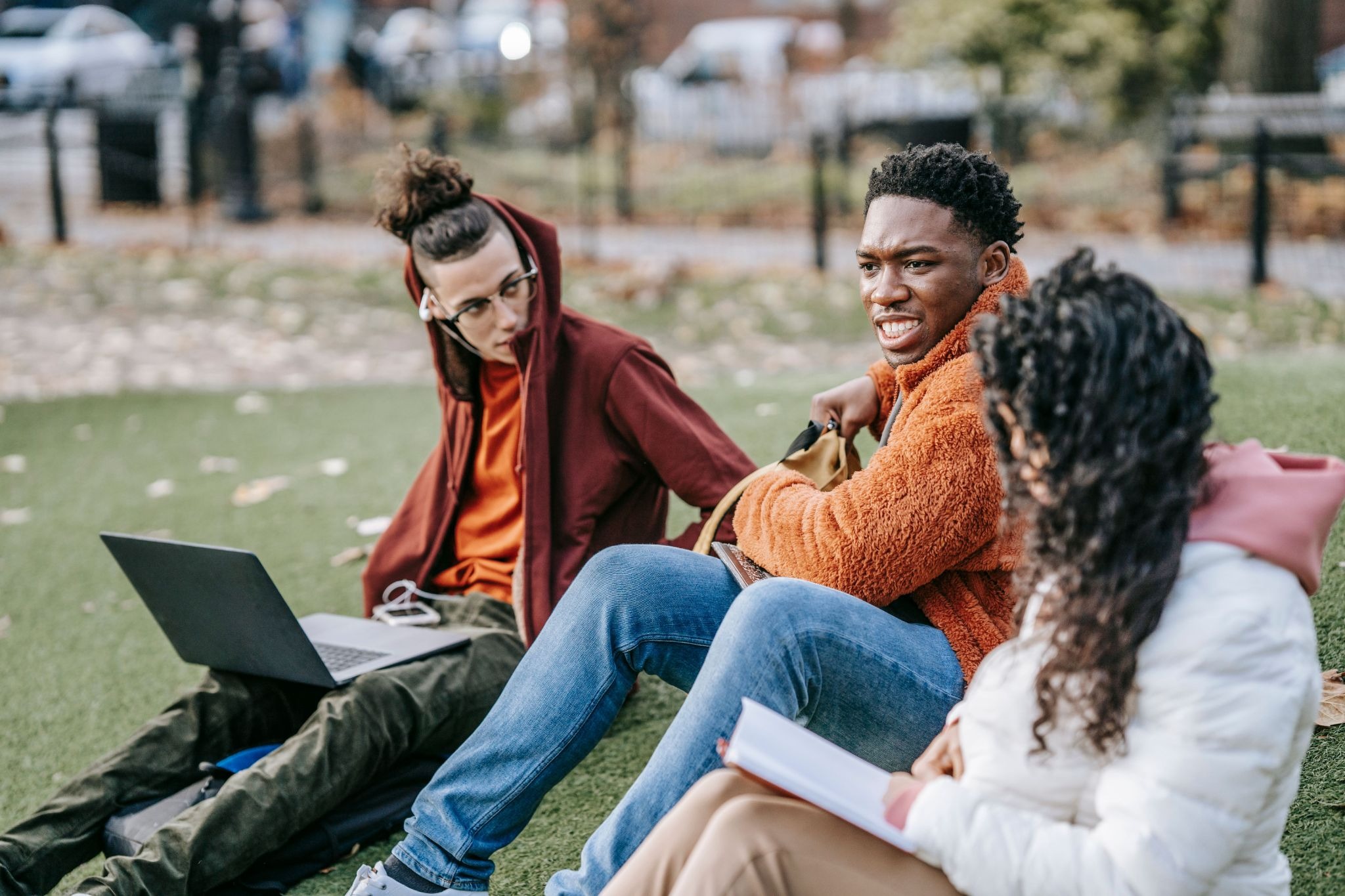 Three students sitting together on an outdoor lawn with books and a laptop, appearing to study while engaged in a serious conversation.