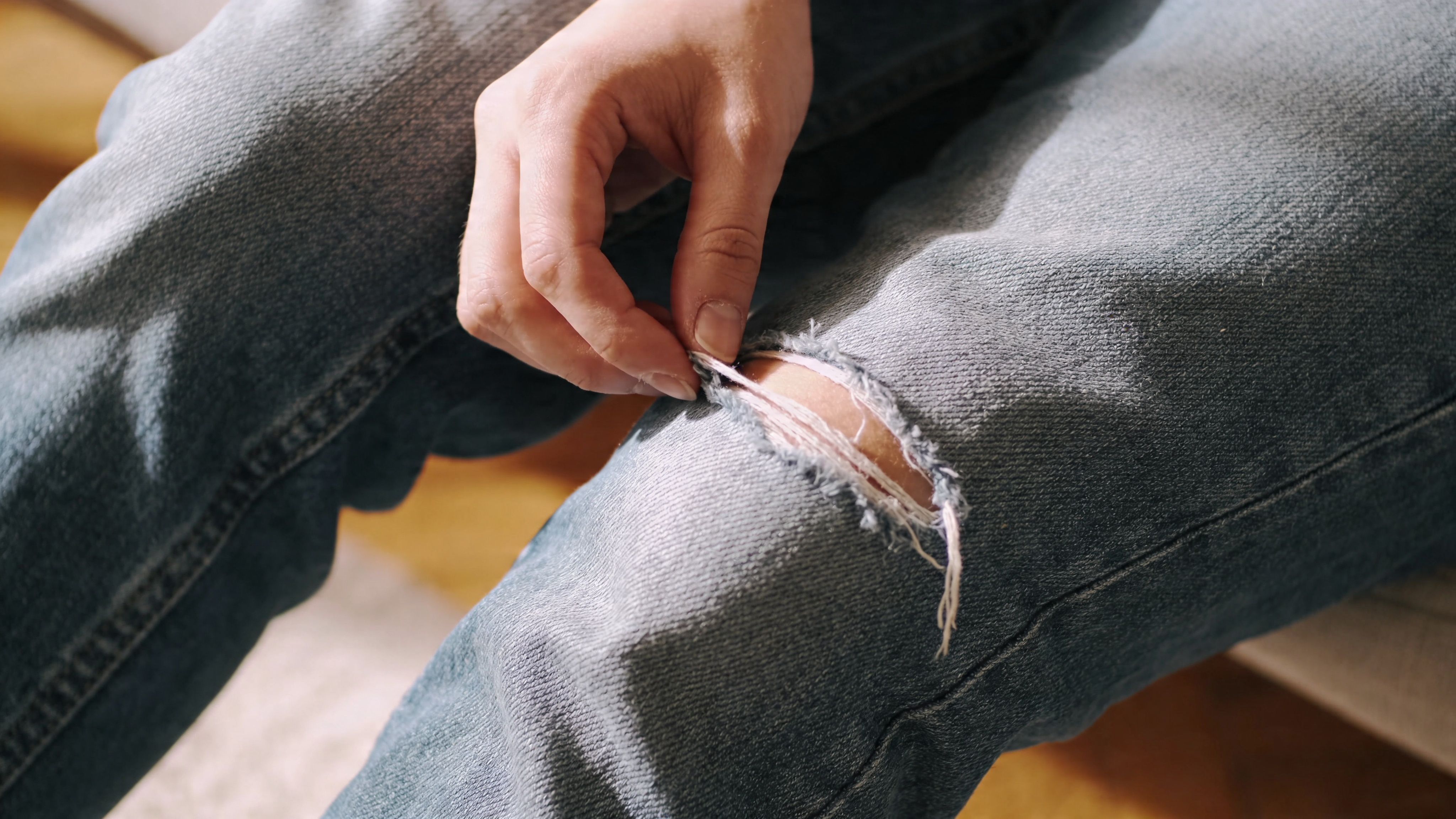 A close-up view of a person using their hand to adjust a rip in their blue jeans