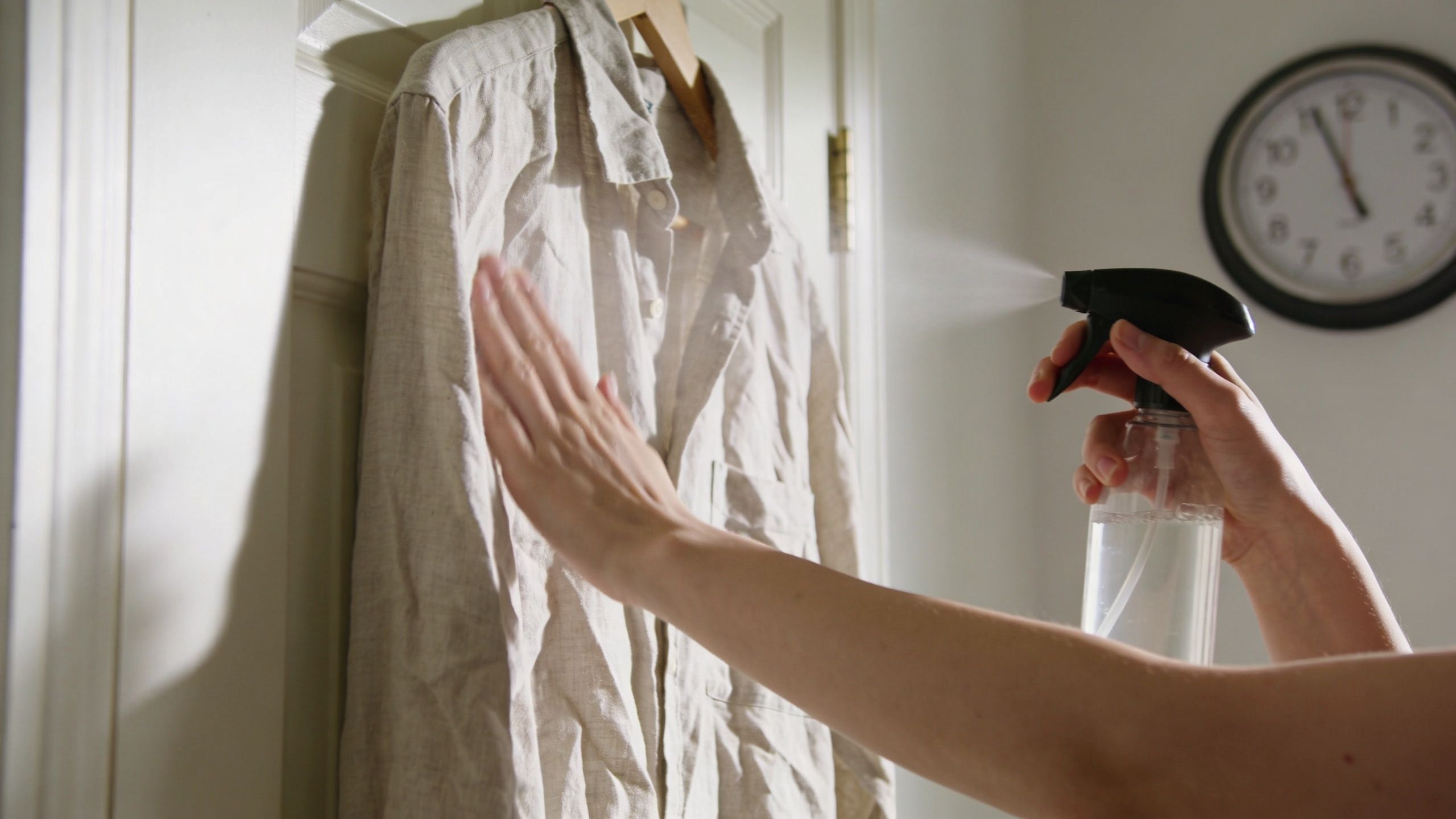 A person using a spray bottle to remove wrinkles from a light-colored linen shirt hanging on a door.