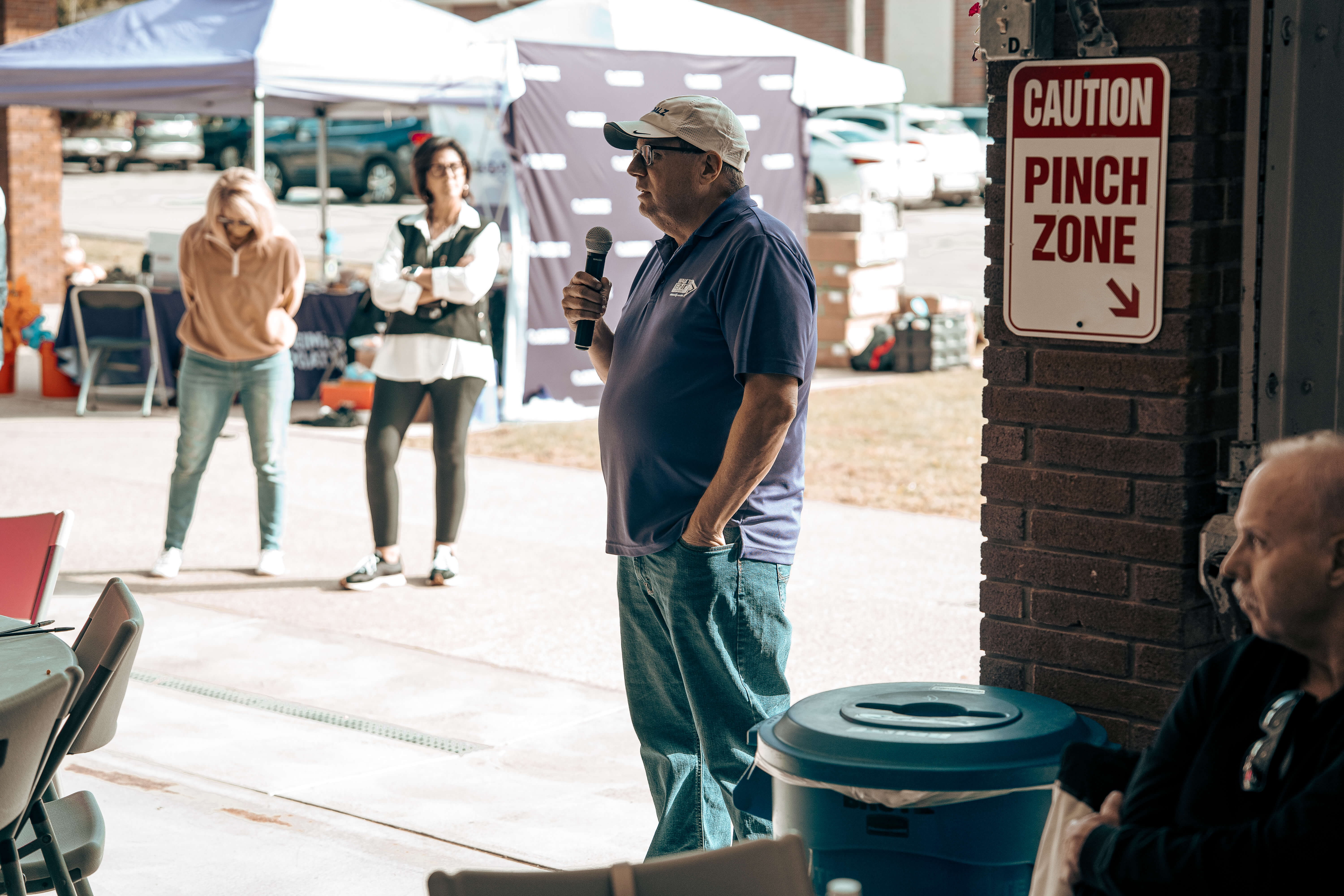 Man speaking with microphone at outdoor event near caution pinch zone sign