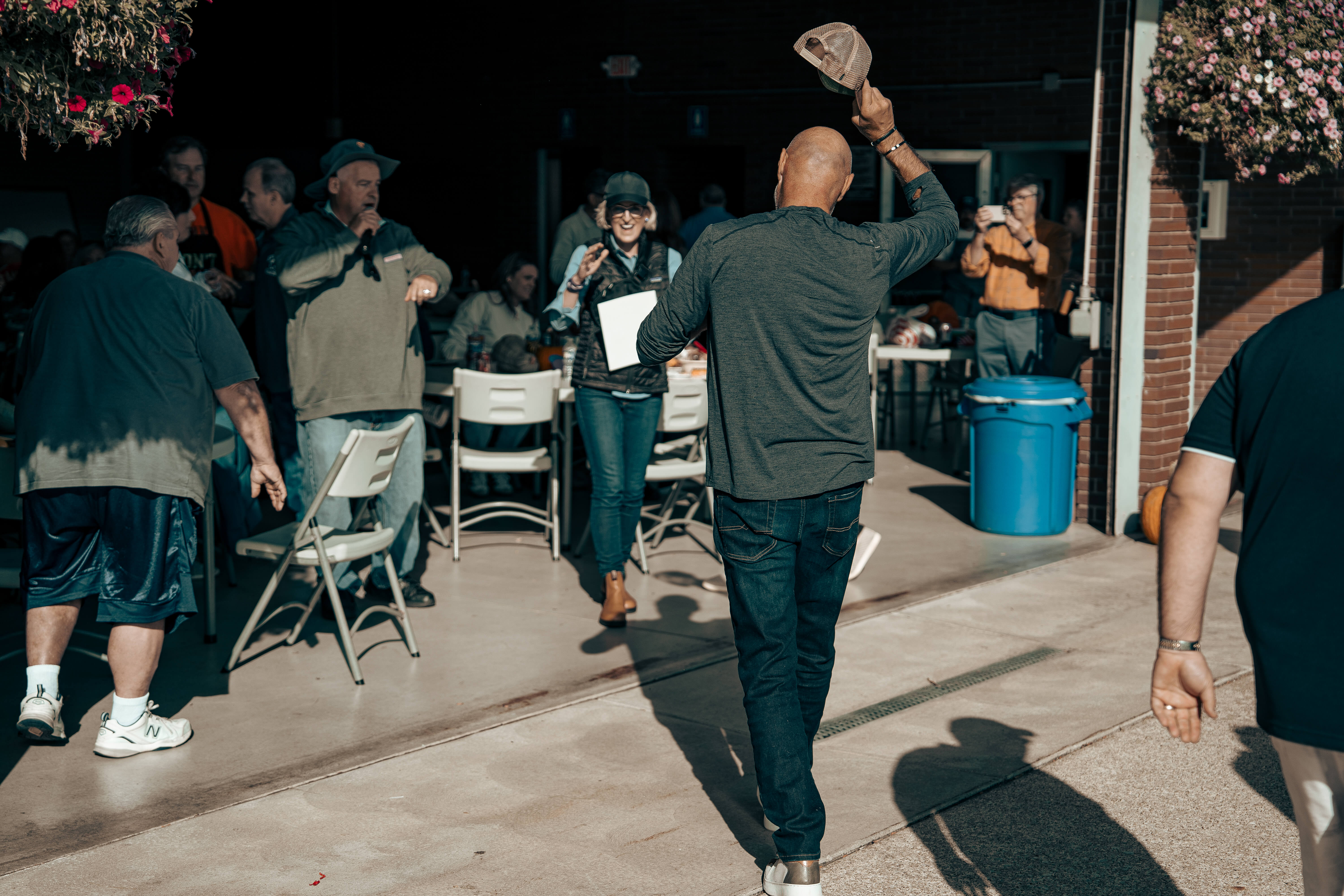 Bald man waving hat at outdoor gathering with chairs and people