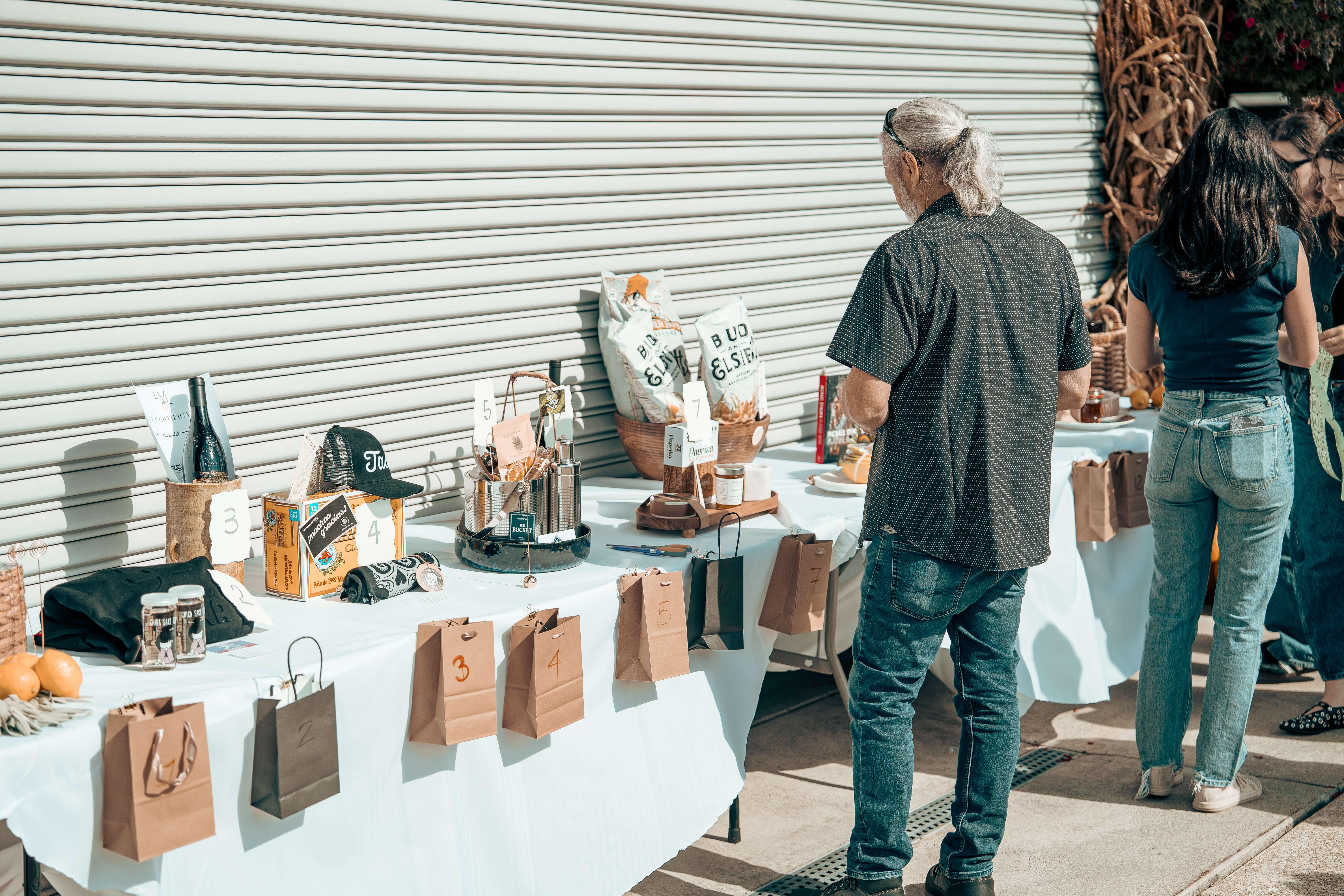 People browsing market stall with numbered brown bags and various products